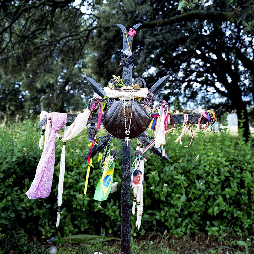 Black metal cross decorated with fabric, ribbons, pearls, a suspended metal cross and a photo of a person, set in a verdant environment with trees in the background.
