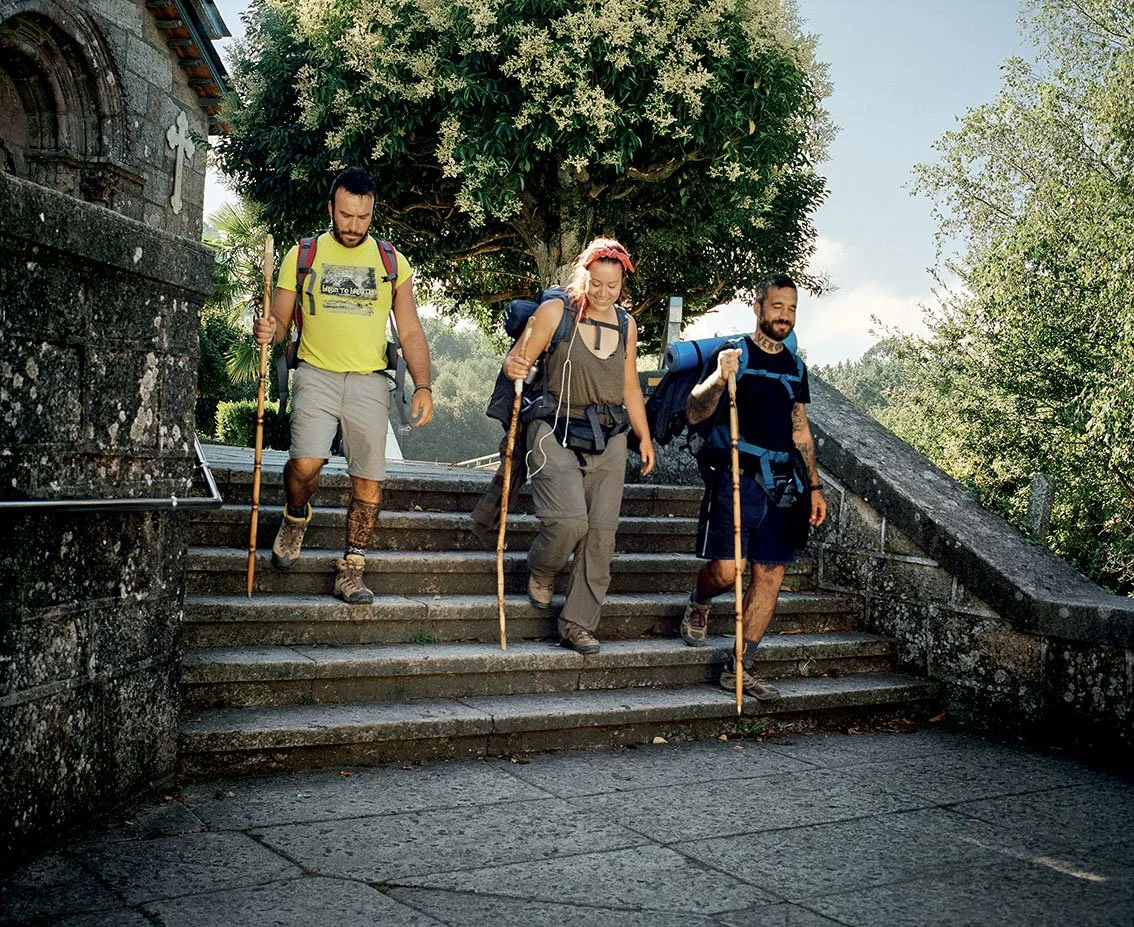 Three people in hiking clothes descend a stone staircase outside, carrying backpacks and walking sticks, under a large tree.