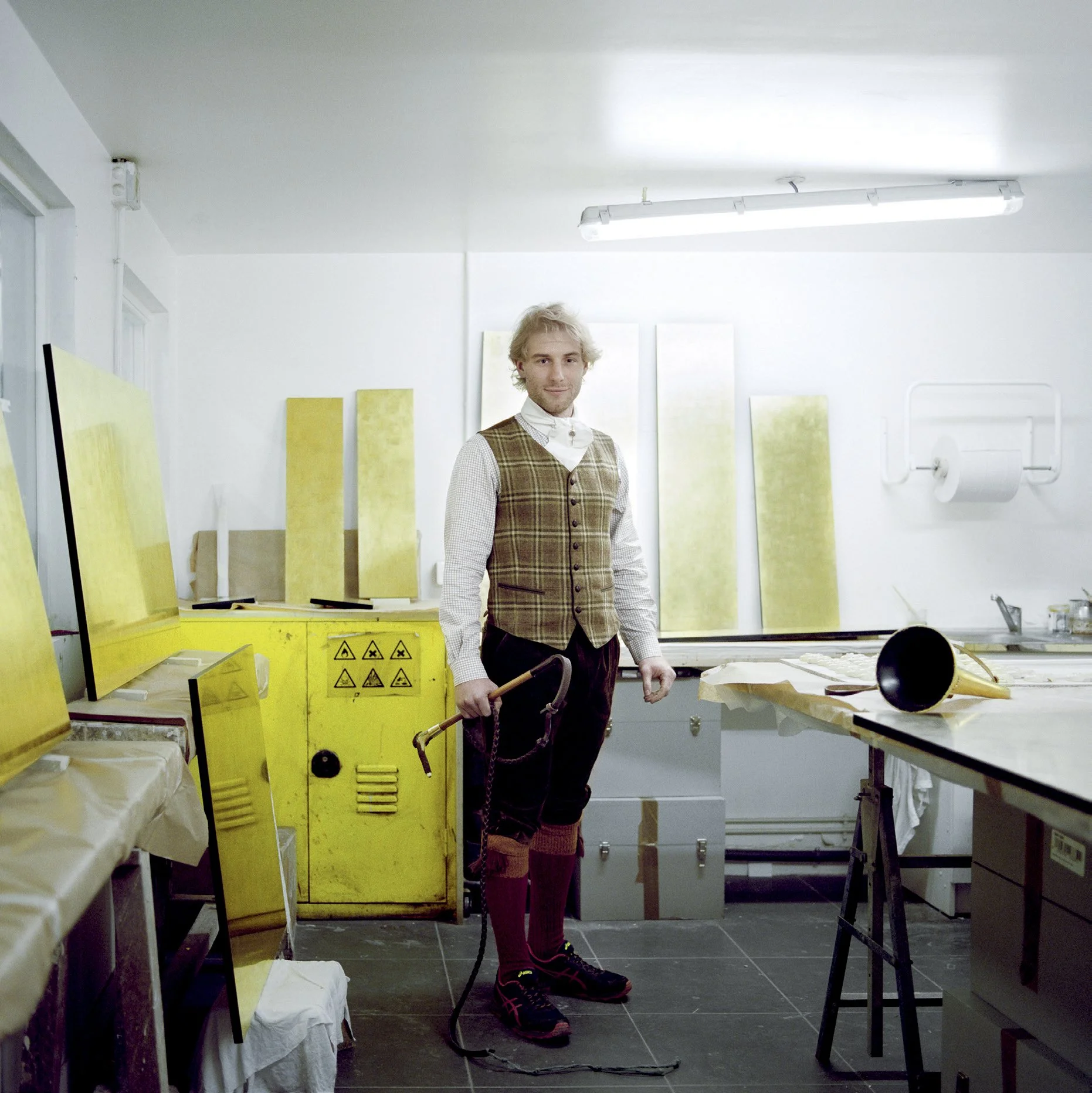 A man dressed in traditional Scottish costume with camera cord in hand, standing in a studio with works of art in progress in a bright environment.