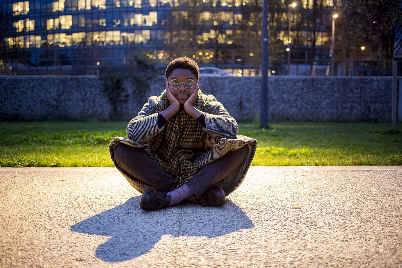 A person sitting cross-legged on the ground, hands over face, in an urbanized area in the evening, with warm lighting and a building in the background.