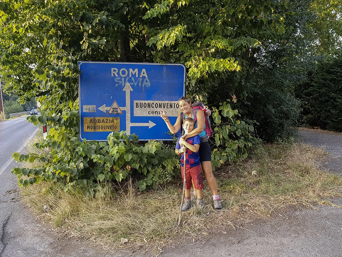 A woman and a boy pose in front of a road sign indicating several directions to Rome, Buonconvento, Abbadia Monteoliveto, with dense vegetation in the background.