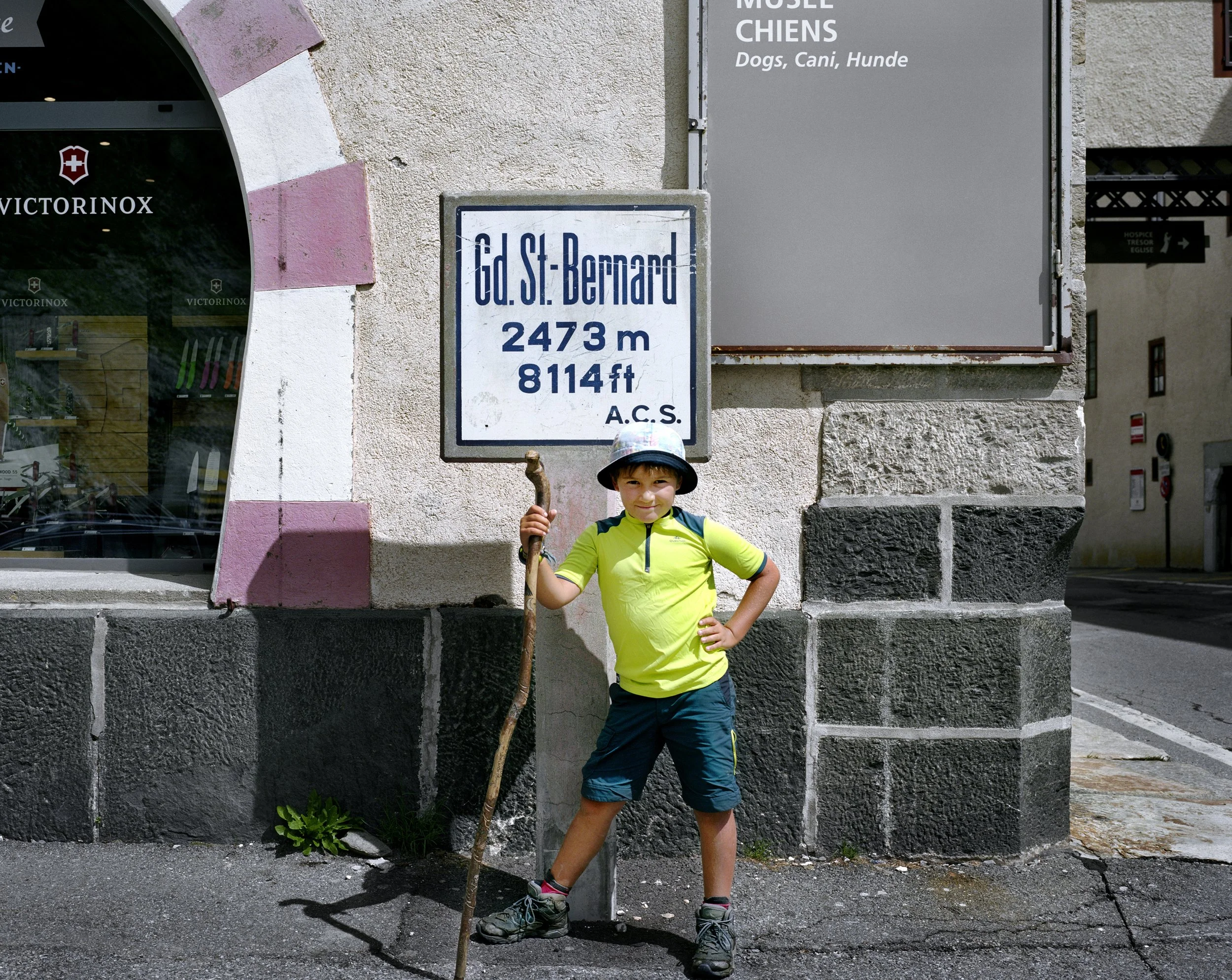 A smiling boy in hiking gear, wearing a hat and holding a stick, stands in front of a plaque indicating the summit of the Gd. St. Bernard mountain at 2473 meters above sea level.