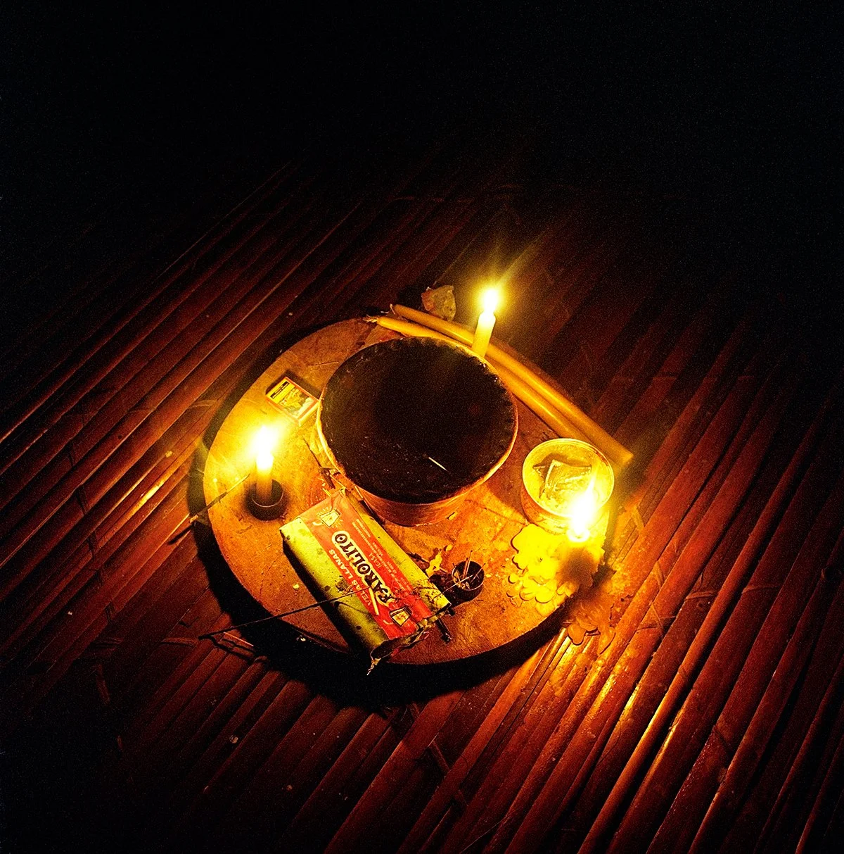 A wooden table with lit candles, a metal pot, a box of matches and a glass. The table is set on a bamboo floor.