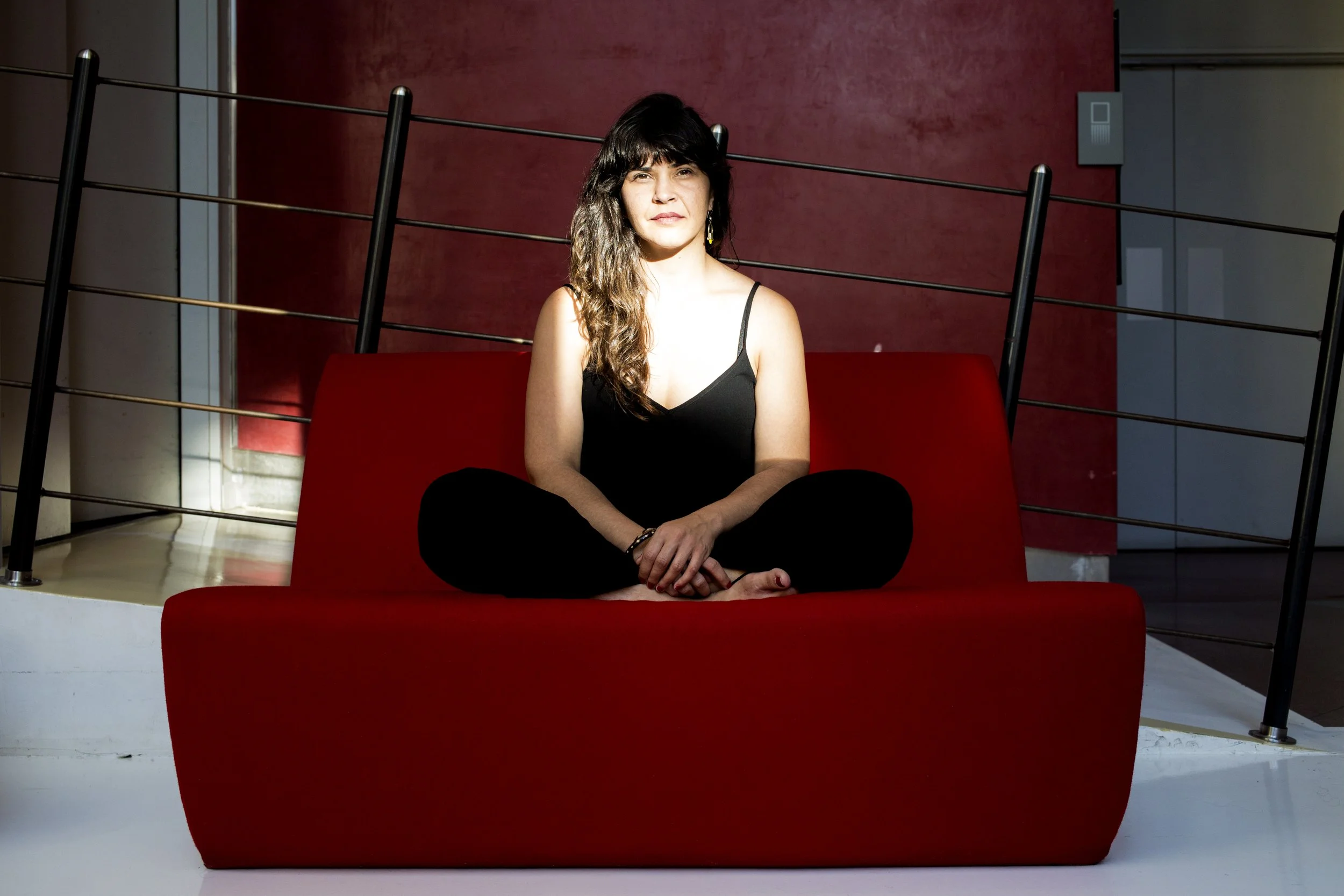 Woman seated cross-legged on a red sofa, wearing black clothing, with a red wall in the background.
