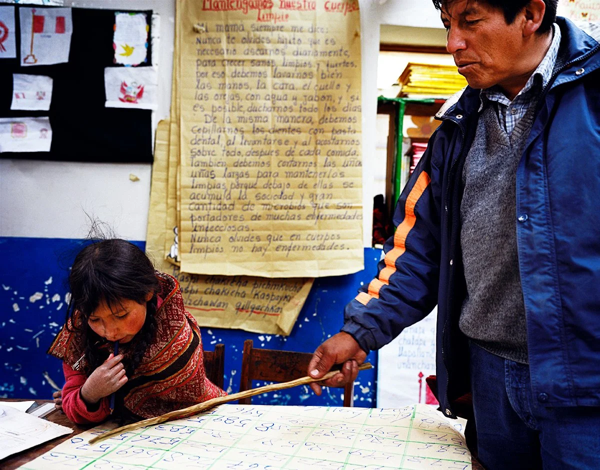 A man shows a young girl how to write with a wooden stick at a table, in a room decorated with posters and papers written in Spanish.