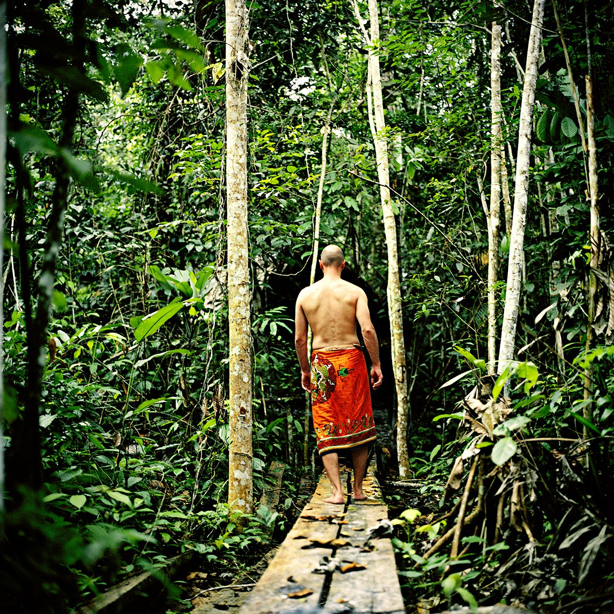 A naked man wearing an orange sarong walks across a wooden bridge in a dense green forest.