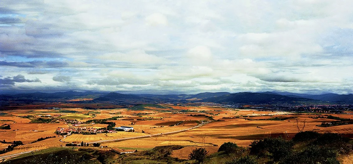 Rural landscape with golden agricultural fields, roads, a few buildings, mountains in background and cloudy sky.