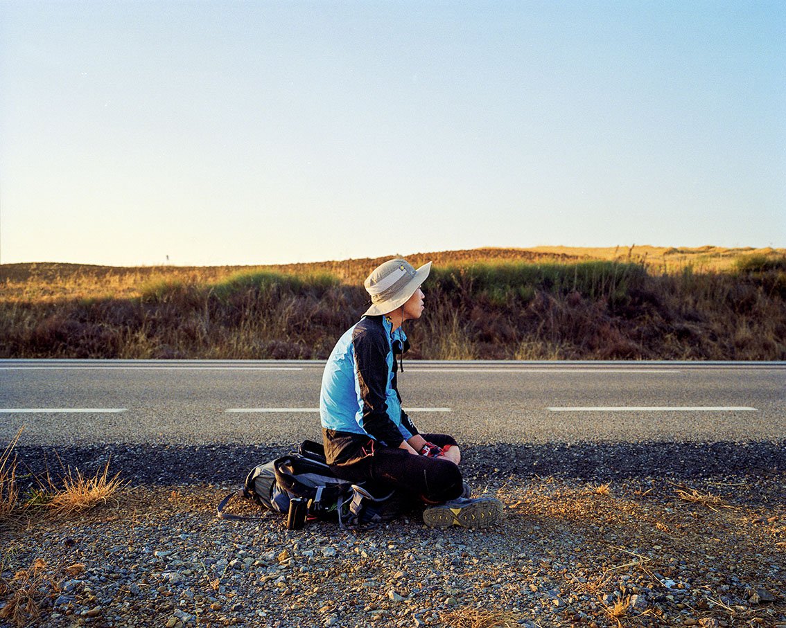 A woman sitting on the side of the road, wearing a hat, a blue and black coat and a rucksack, gazing towards the horizon at sunset in a desert landscape.