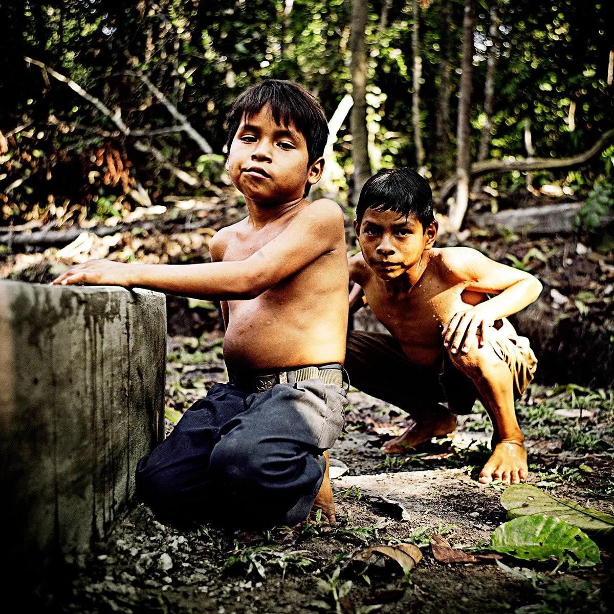 Two naked boys in a dense forest, one crouched near a concrete structure and the other picked up off the ground, both looking at the lens.