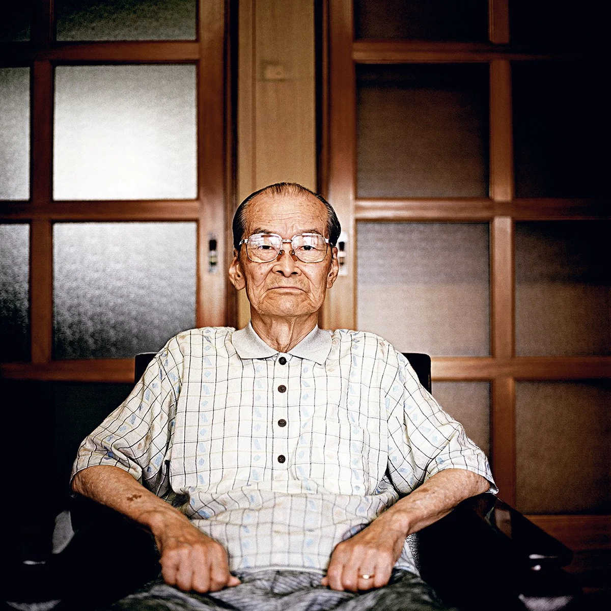 Un homme âgé avec des lunettes, portant une chemise à carreaux, assis dans une pièce avec un fond en bois et des panneaux en Micha. Il regarde directement l'appareil photo avec une expression sérieuse.