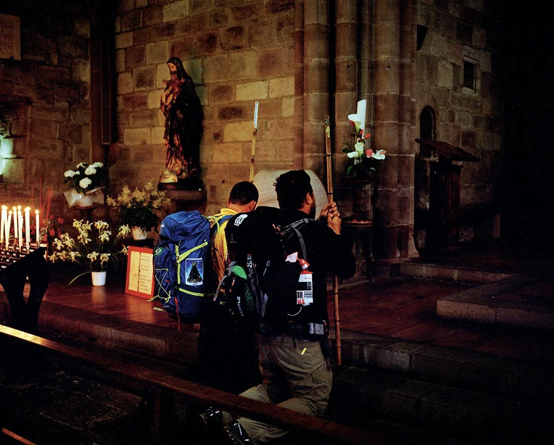 Two men carrying backpacks and walking sticks, pray in a church in front of a religious statue, flowers and lit candles.