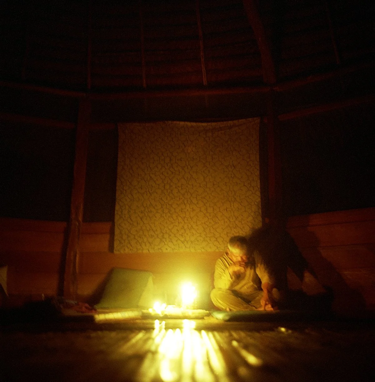 A person sitting in a dark room, lit only by a burning candle, with wooden walls and a yellow and orange carpet.