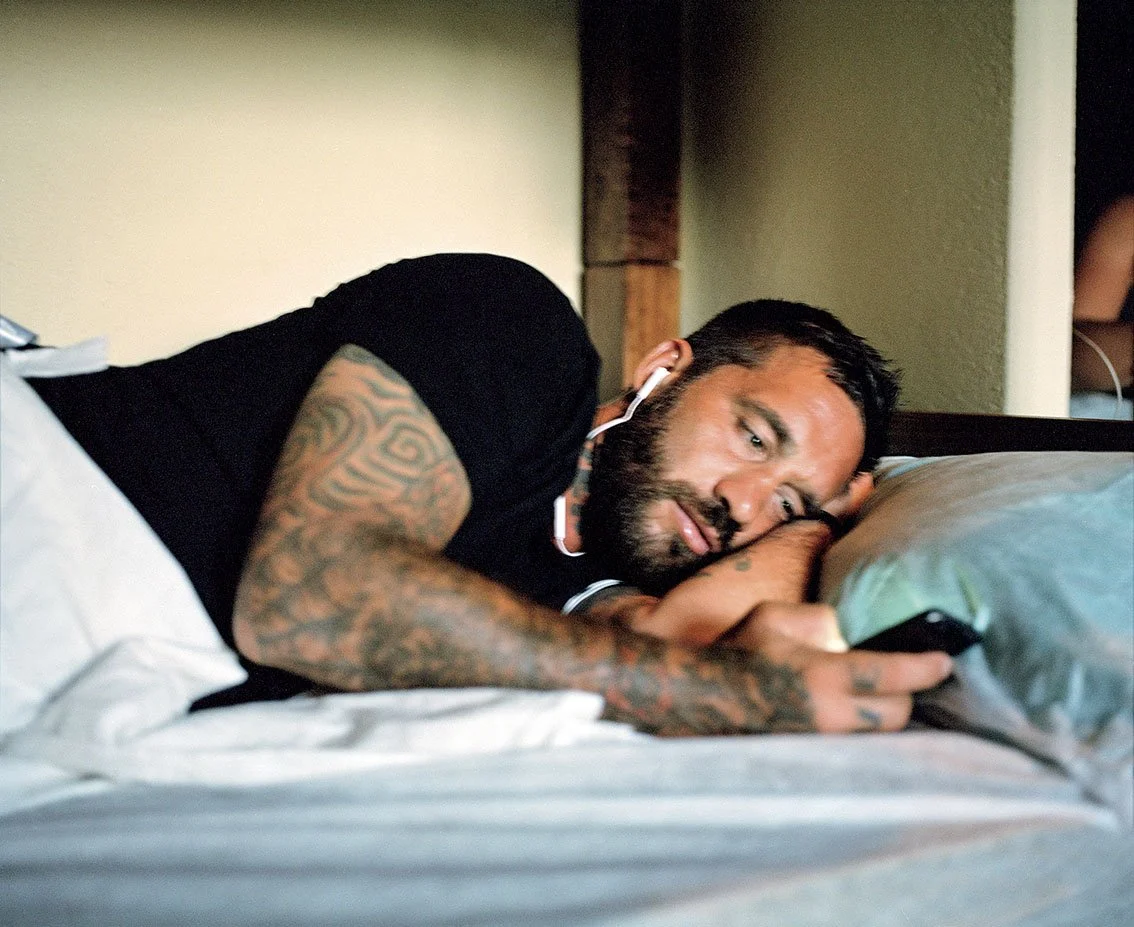 A man with tattoos resting on a bed, using a phone with headphones.