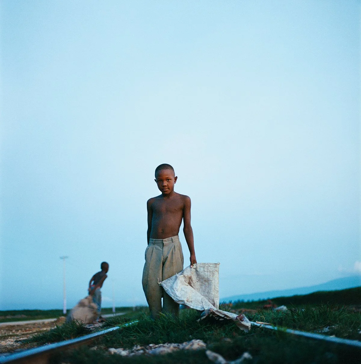 A naked boy in beige pants, holding a flag or cloth, stands on a vacant lot during sunset. In the background, another child is present. The sky is clear with shades of blue.