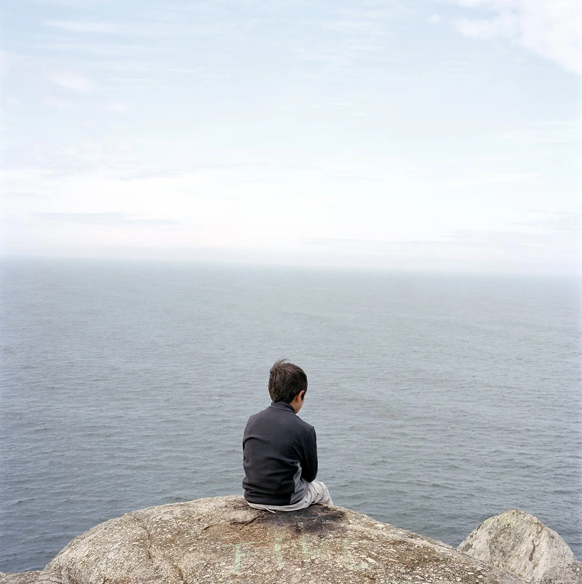 A boy sitting on a rock, looking out at the ocean in the distance, in a coastal setting.