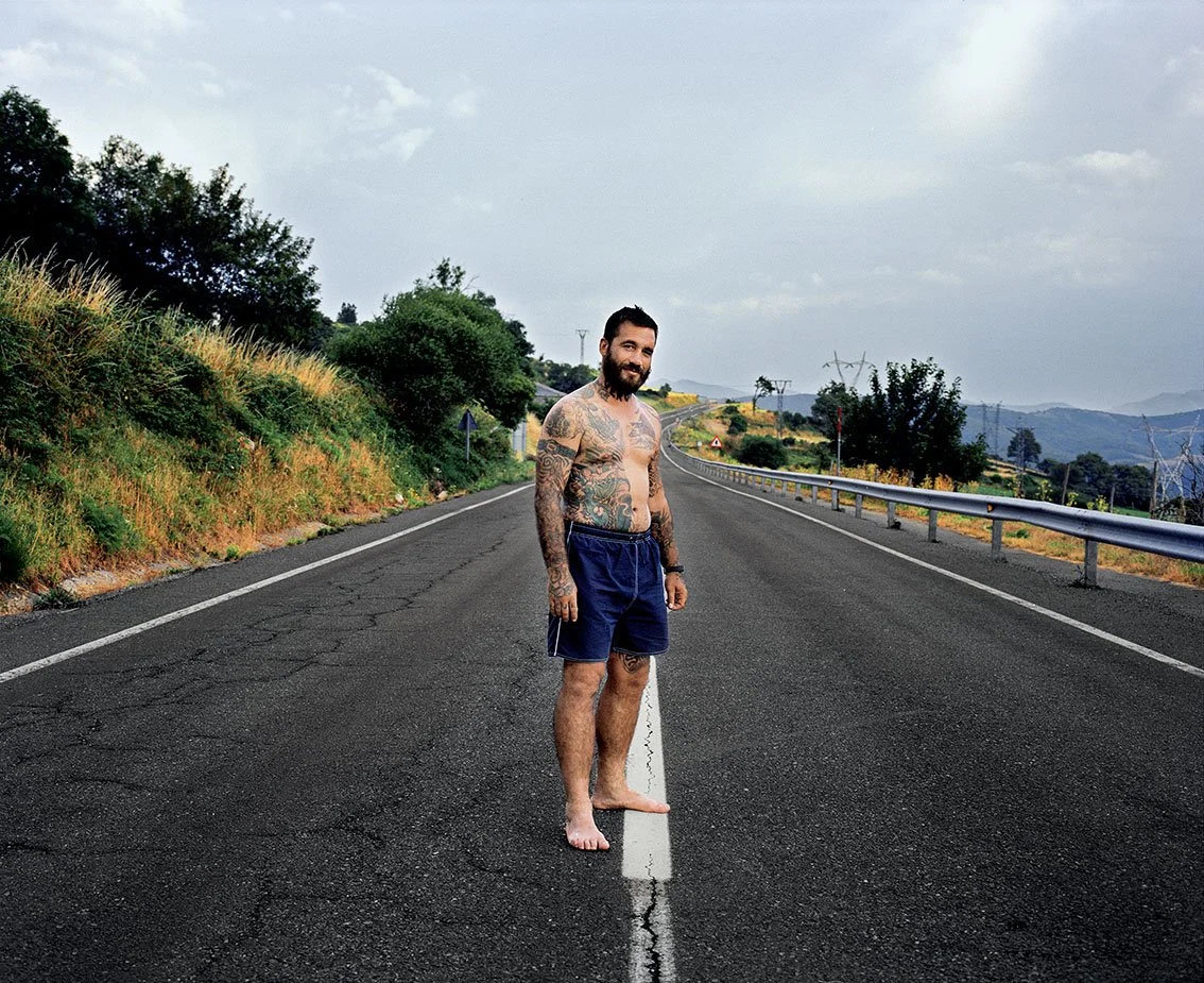 A tattooed man in shorts, standing barefoot on the center line of a country road, surrounded by hills and trees, under a cloudy sky.