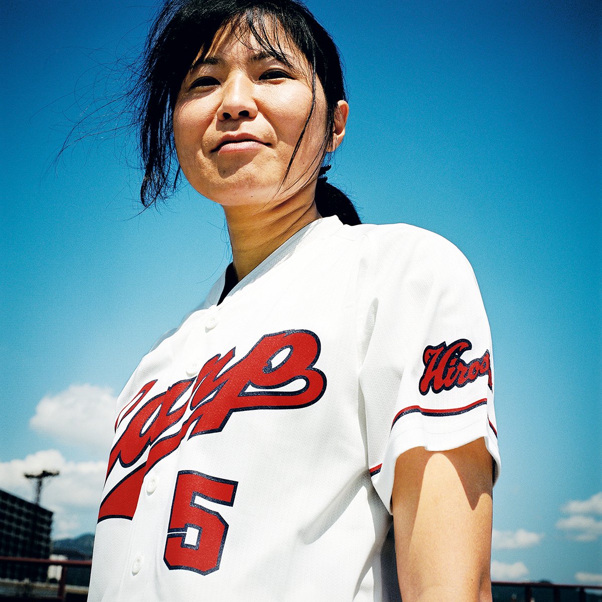 Femme portant un maillot de baseball blanc avec le mot 'Hiroshima' en rouge, sous un ciel bleu clair