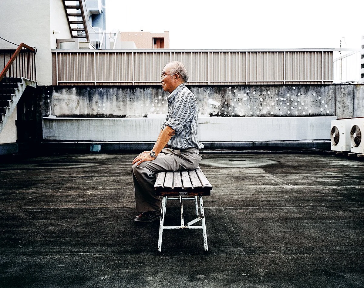 Un homme âgé assis seul sur un banc en bois sur le toit d'un bâtiment urbaine. L'arrière-plan montre un mur en béton et des appareils de climatisation.