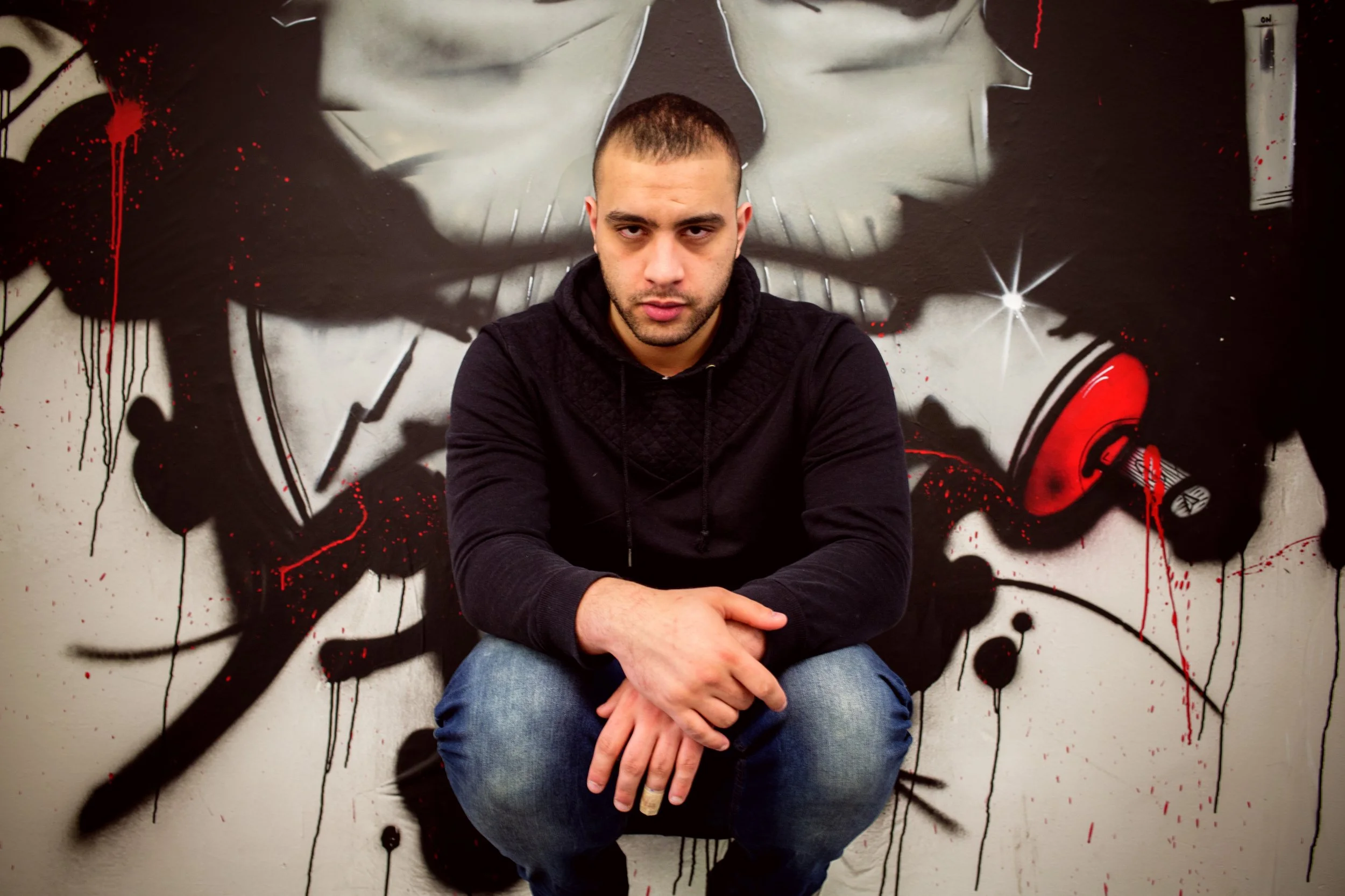 Young man seated in front of a graffiti wall depicting a face in black and white with stylized elements in red and black.