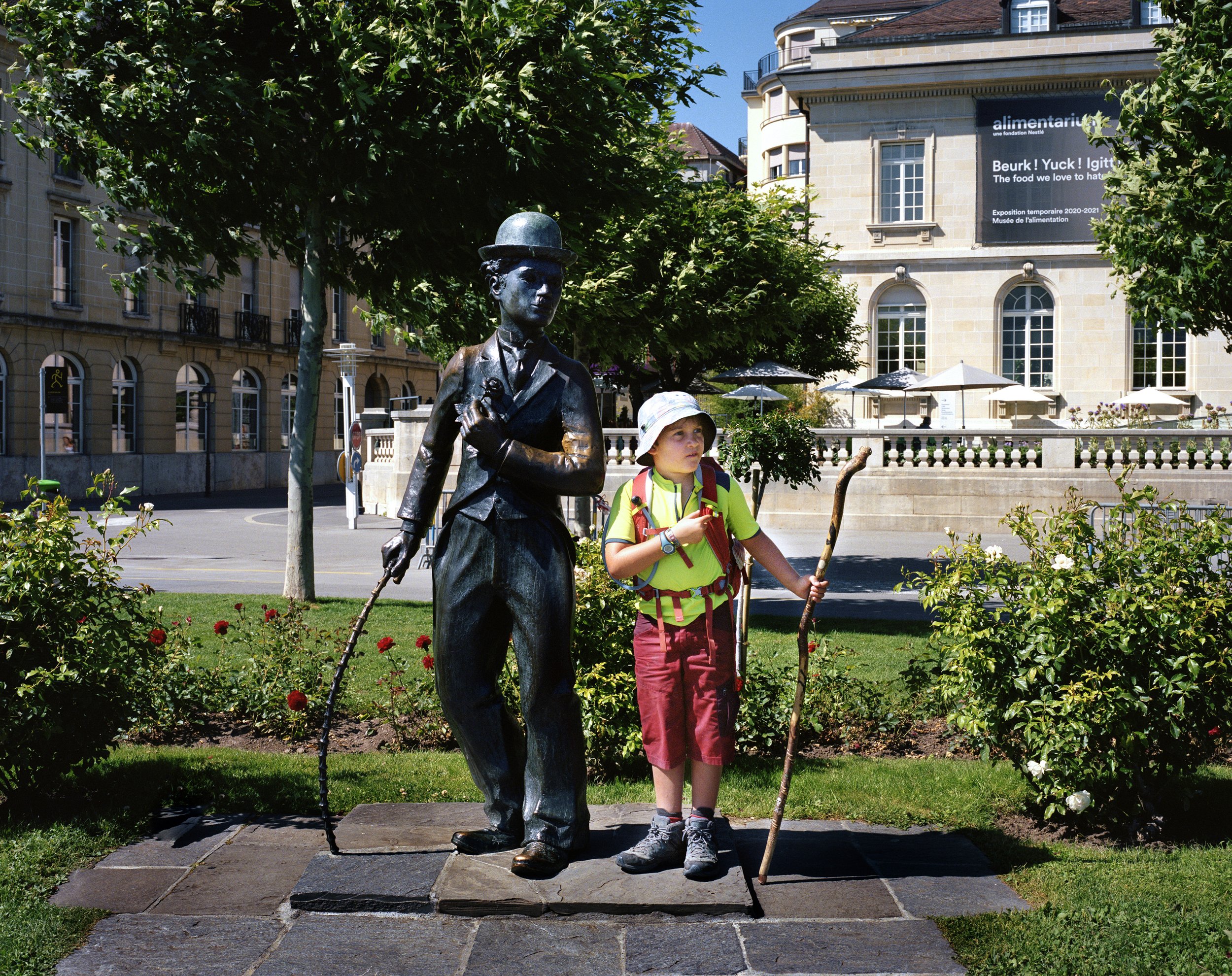 A young boy in a green shirt, red shorts and white hat, carrying a backpack, stands next to a bronze statue of an old-fashioned woman in an urban park. The statue shows a woman wearing a hat and a traditional costume.