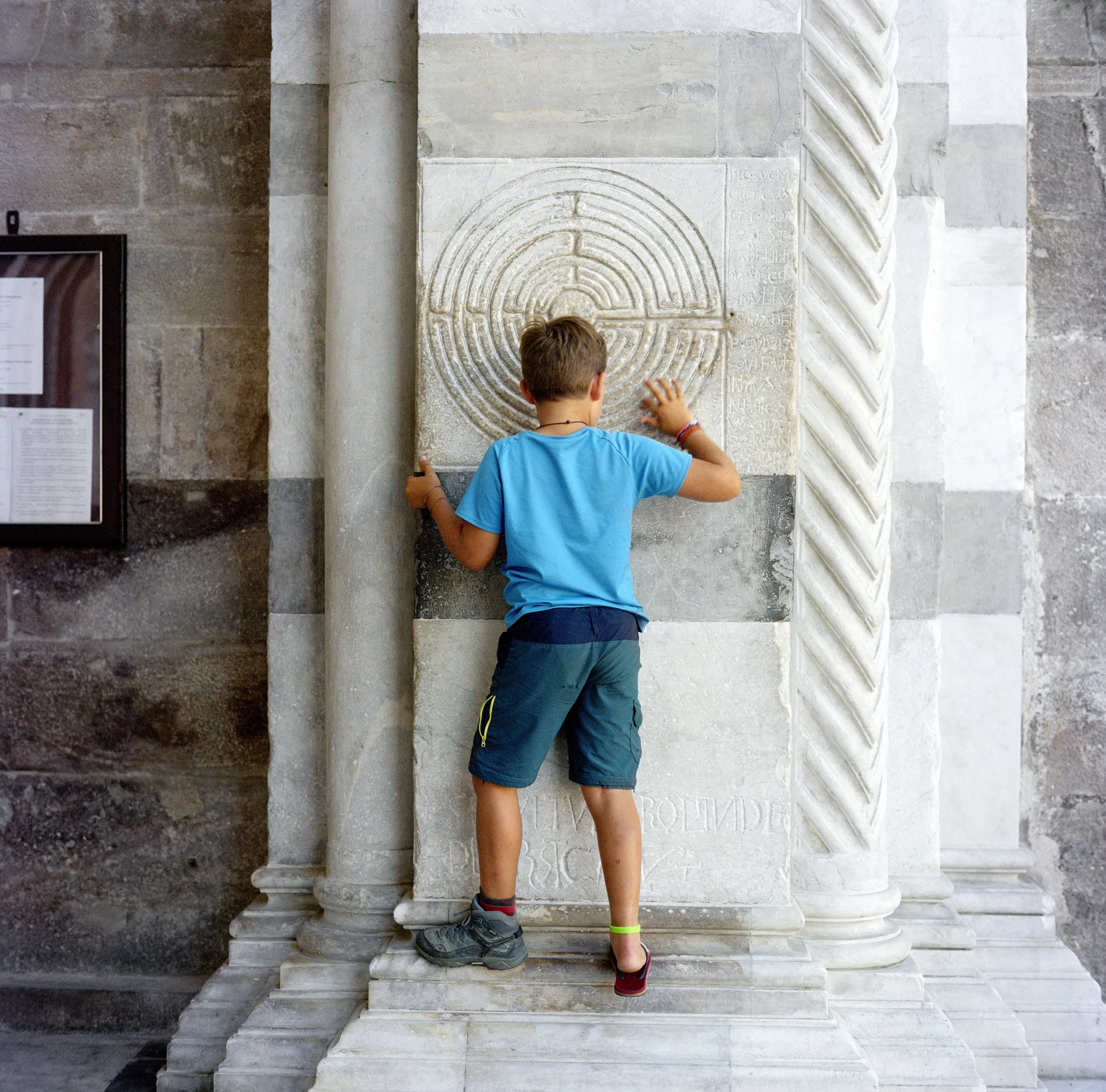 Young boy in blue T-shirt and shorts sitting in front of a fresco or stone monument in a museum.