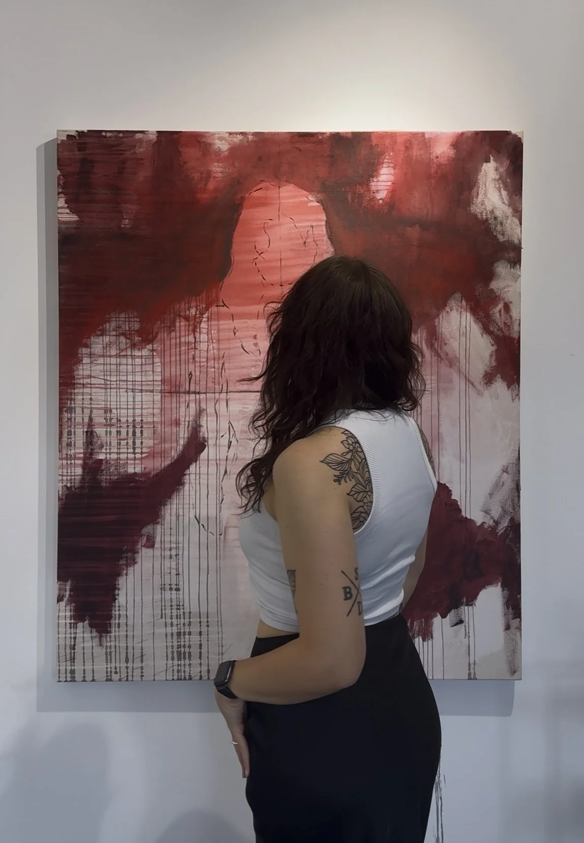 Eine Frau mit dunklem, lockigem Haar trägt ein weißes Top und eine schwarze Hose, steht vor einem Kunstwerk mit roten und weißen Farbverläufen, das eine abstrakte, gesichtsloses Figur zeigt.