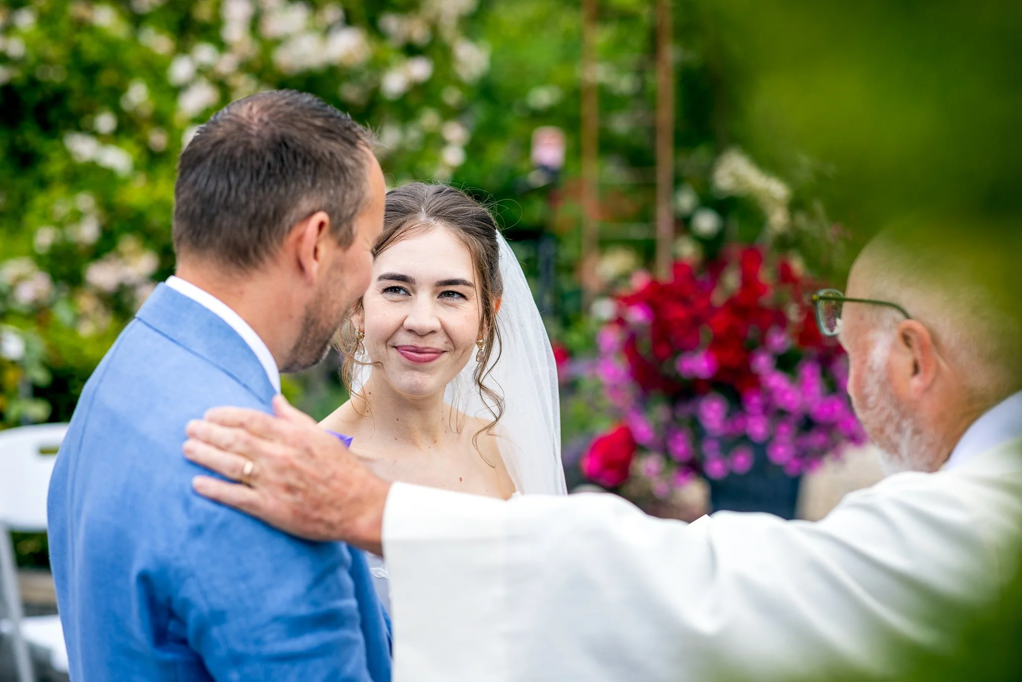 En bryllupsceremoni med en brud og en brudgom som omfavner hverandre, omgitt av fargerike blomster i en utendørs hage, med en seremonileder som holder en tale.