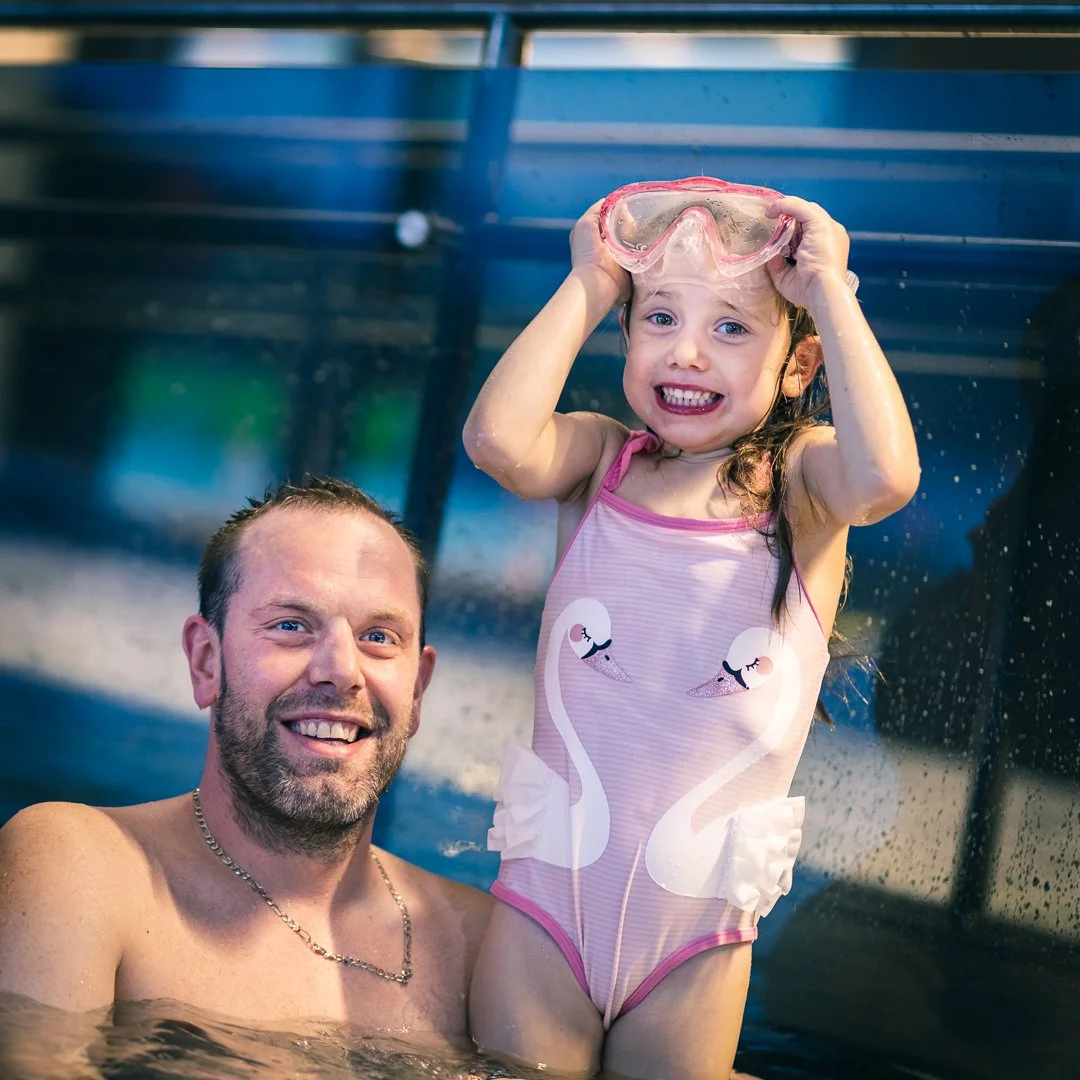 En glad liten jente i badeklær med svaner på, som holder på badebrillene sine mens hun sitter på en voksen manns skuldre i en basseng. Mannen smiler.