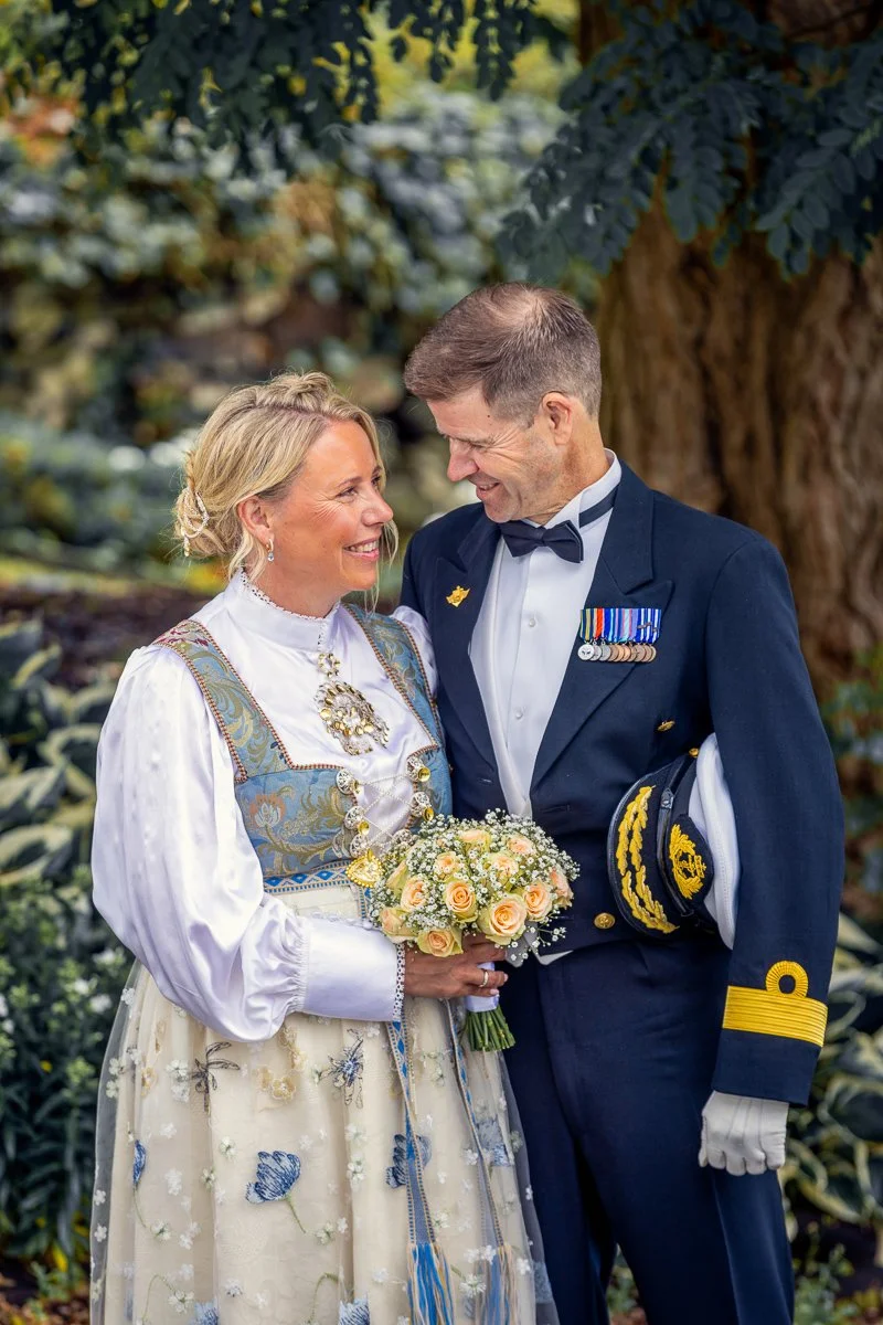 Et bryllupsbilde av et par i tradisjonelle klær, hvor kvinnen holder en bukett gule roser og mannen er i militæruniform, står under et tre.