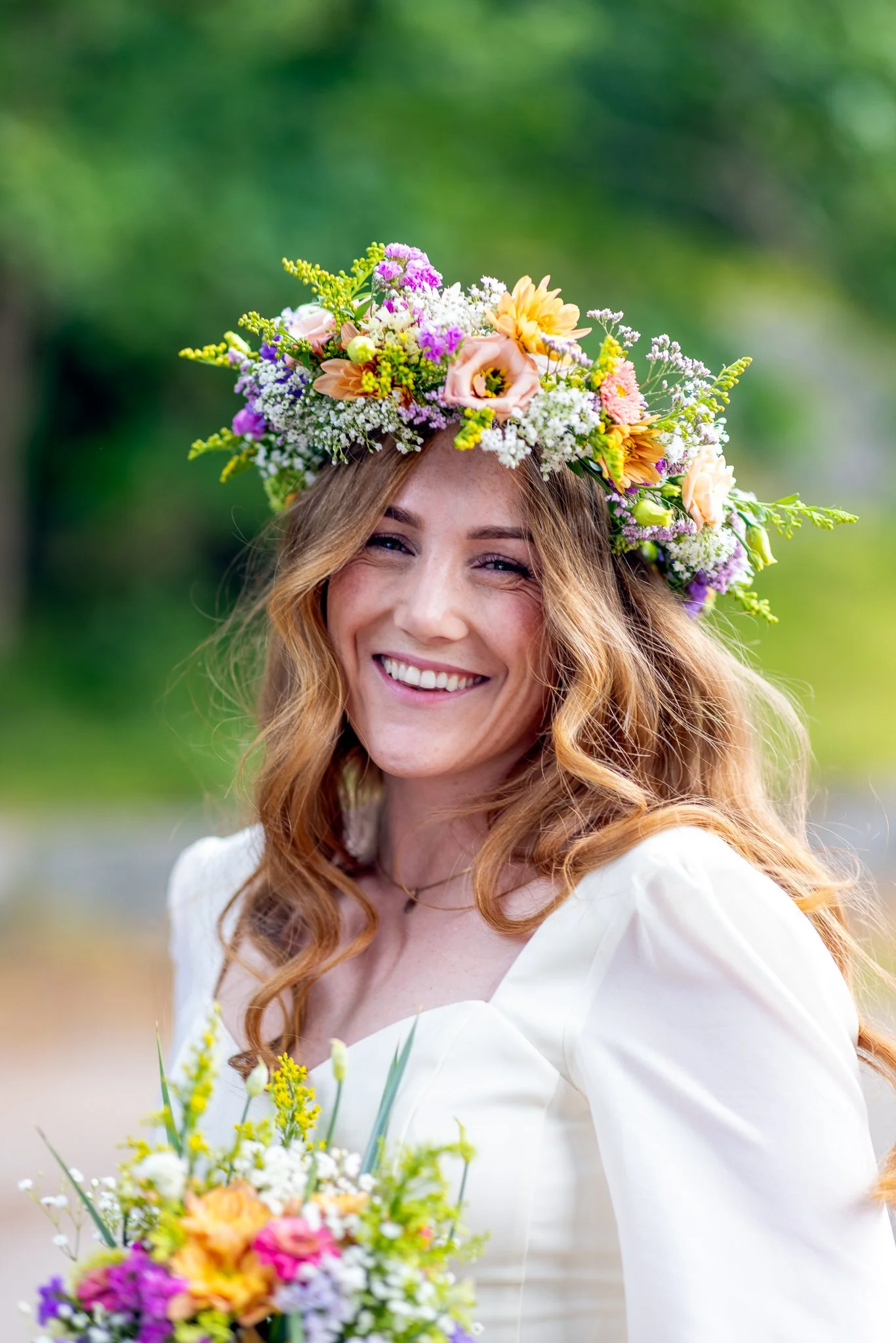 En smilende kvinne med lang, krøllete, rødbrun hår, iført hvit kjole, med en blomsterkrone av ulike fargede blomster på hodet, står utendørs med grønn bakgrunn.