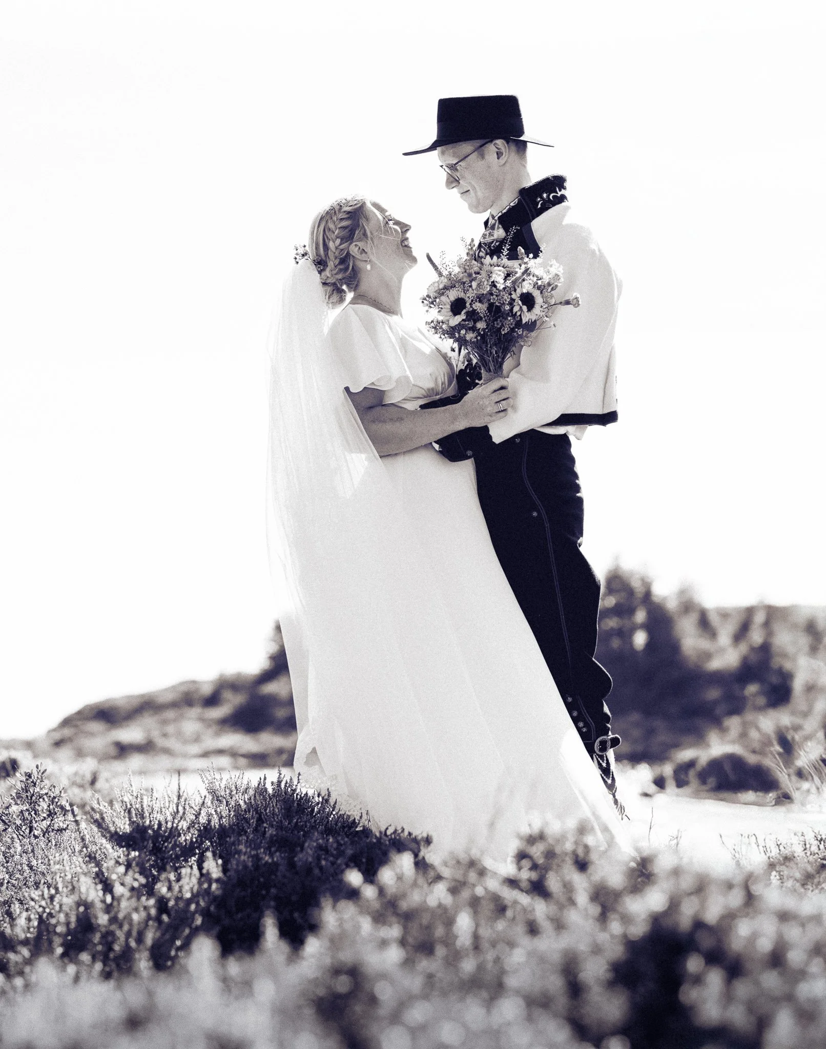 Et bryllupsbilde av et ektepar, kvinnen i brudekjole og mann i tradisjonell folkedrakt, som står på en strand, holder blomster, og ser på hverandre.