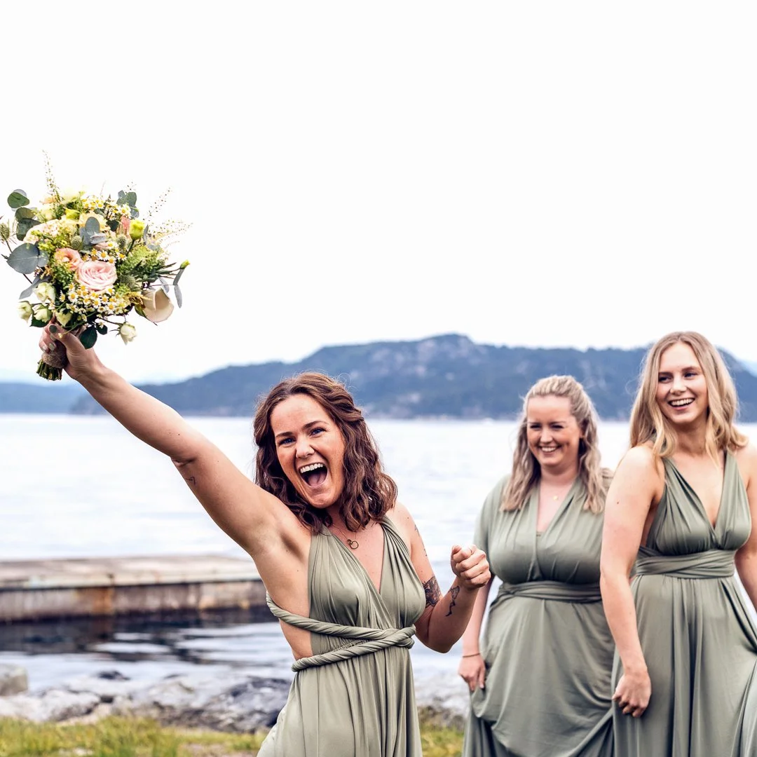 Tre kvinnelige bryllupsdamer i matchende grønne kjoler smiler ved en sjø, en av dem holder opp en blomsterbuket i lufta.