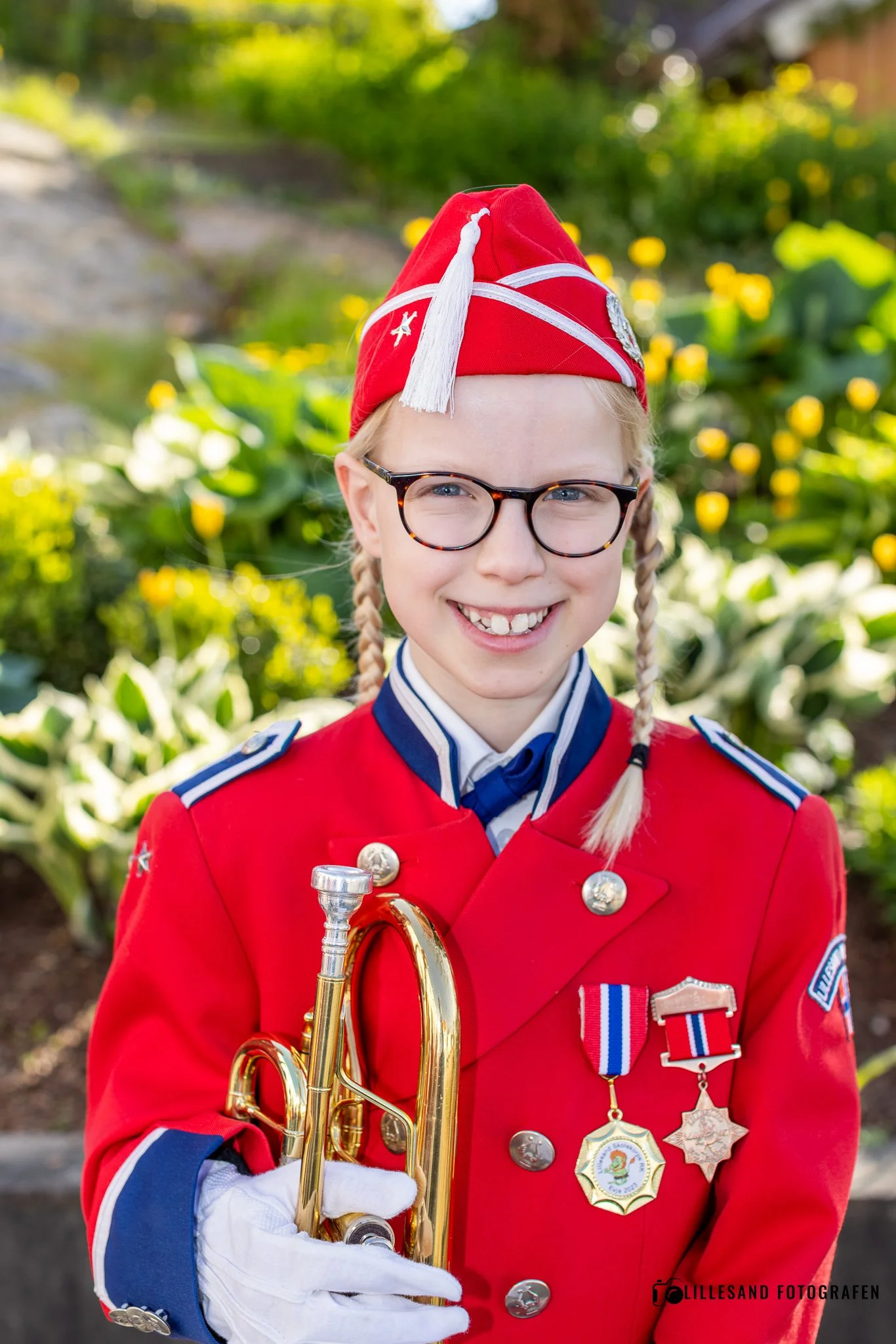 En ung jente iført uniform med medaljer, rød hatt og briller, holder en trompet og smiler. Bakgrunnen er grønn med planter og blomster.