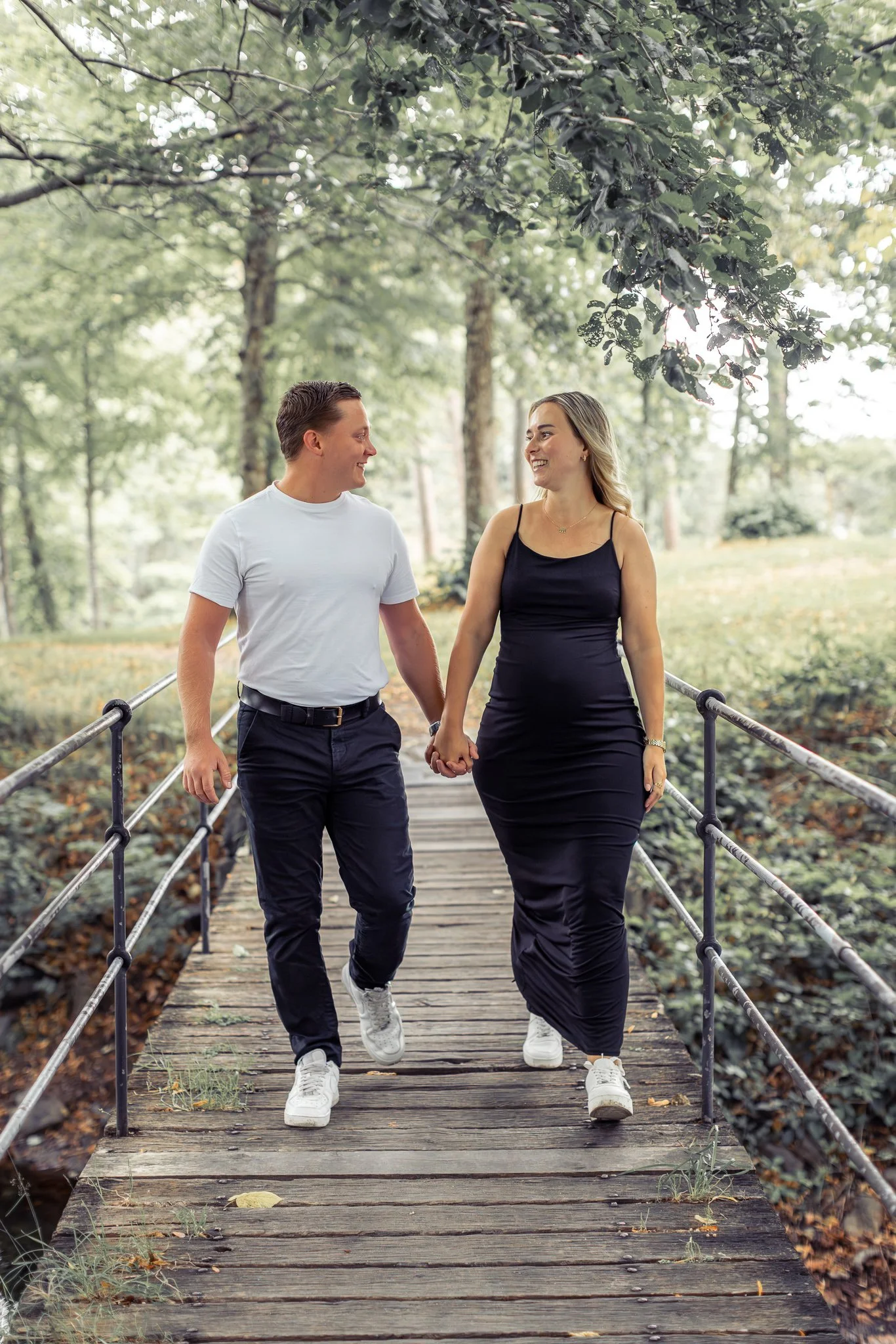 En ung mann og en ung kvinne går hånd i hånd på en trebro i en grønn park, smiler og ser på hverandre.