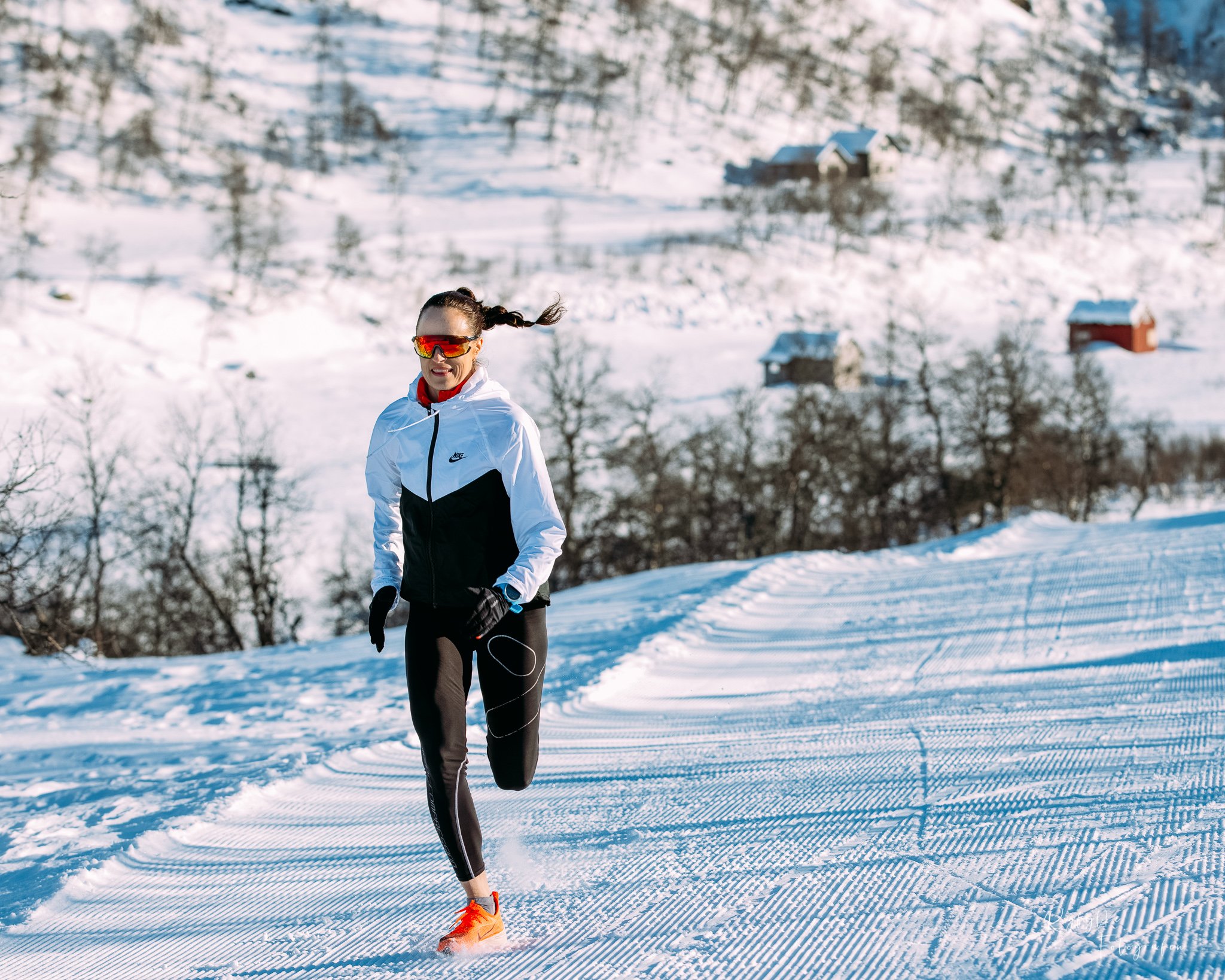En kvinne løper på en snøryggbanen i et snødekt landskap med hus og trær i bakgrunnen, iført sportsutstyr og solbriller.