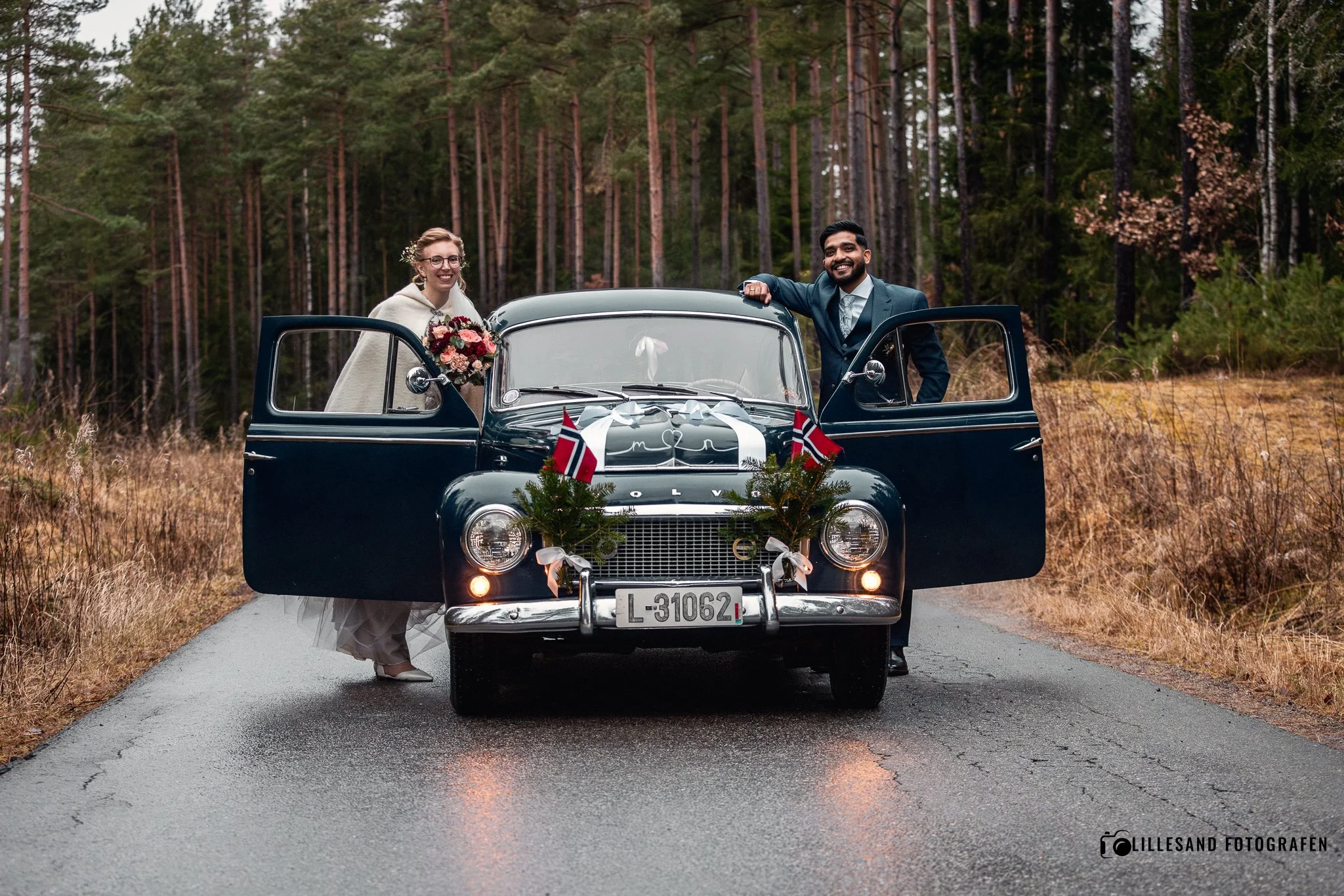 Et ektepar står ved en svart vintagebil med dekorasjoner, på en grusvei i en skog. De ser glade ut, og kvinnen holder en blomsterbukett. Bilen er pyntet med små norske flagg og grønne grener. Dette ser ut til å være en bryllupsfeiring.