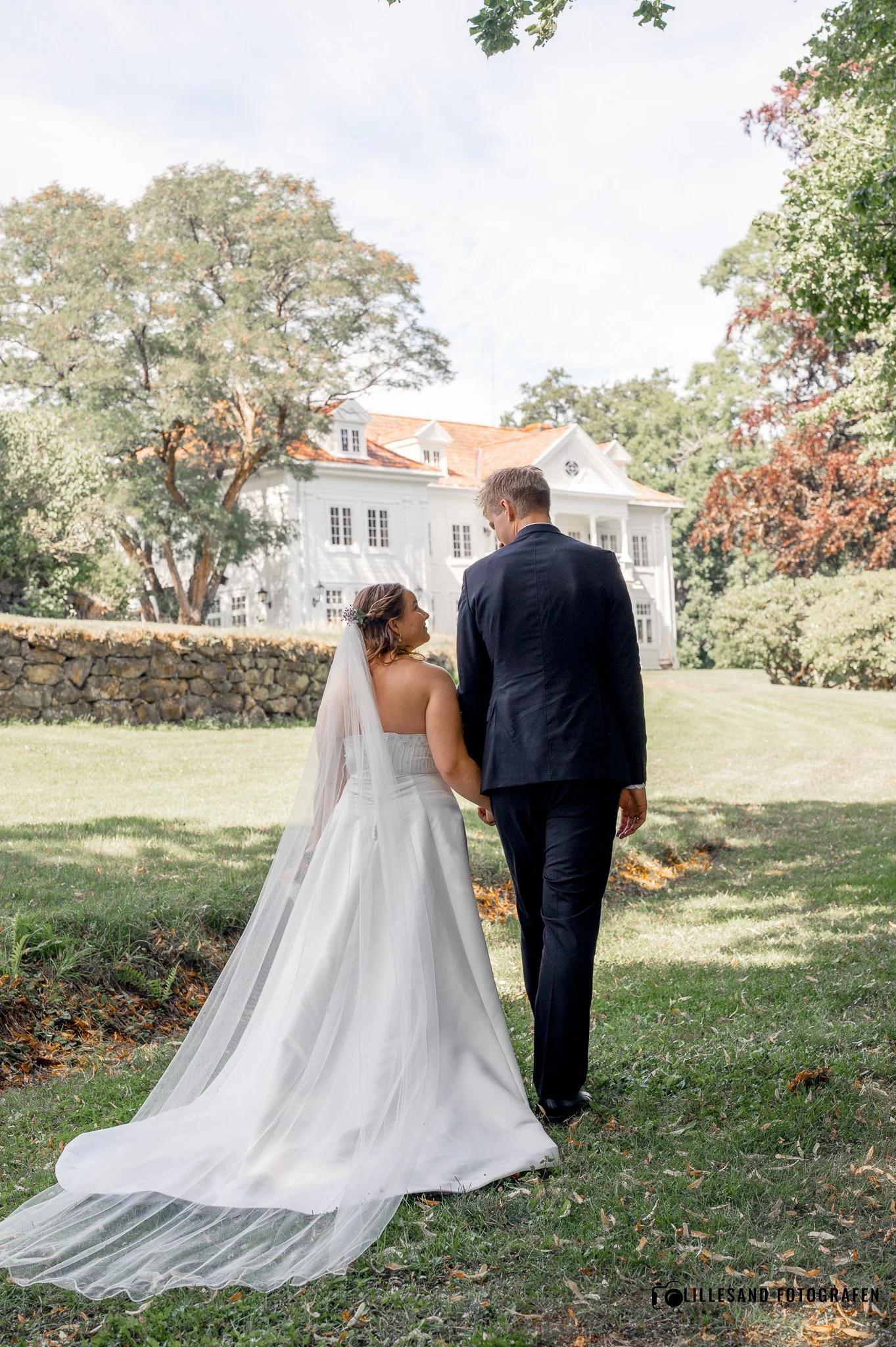 En brud og brudgom går hånd i hånd på en grønn gresslette foran ethvitt hus, omgitt av trær med grønne og brune blader, i et lyst solskinn.