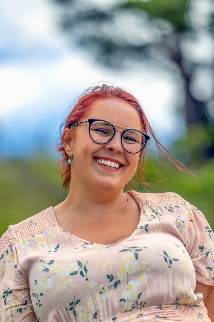 En kvinne med rødt hår og briller smiler i en utendørs setting