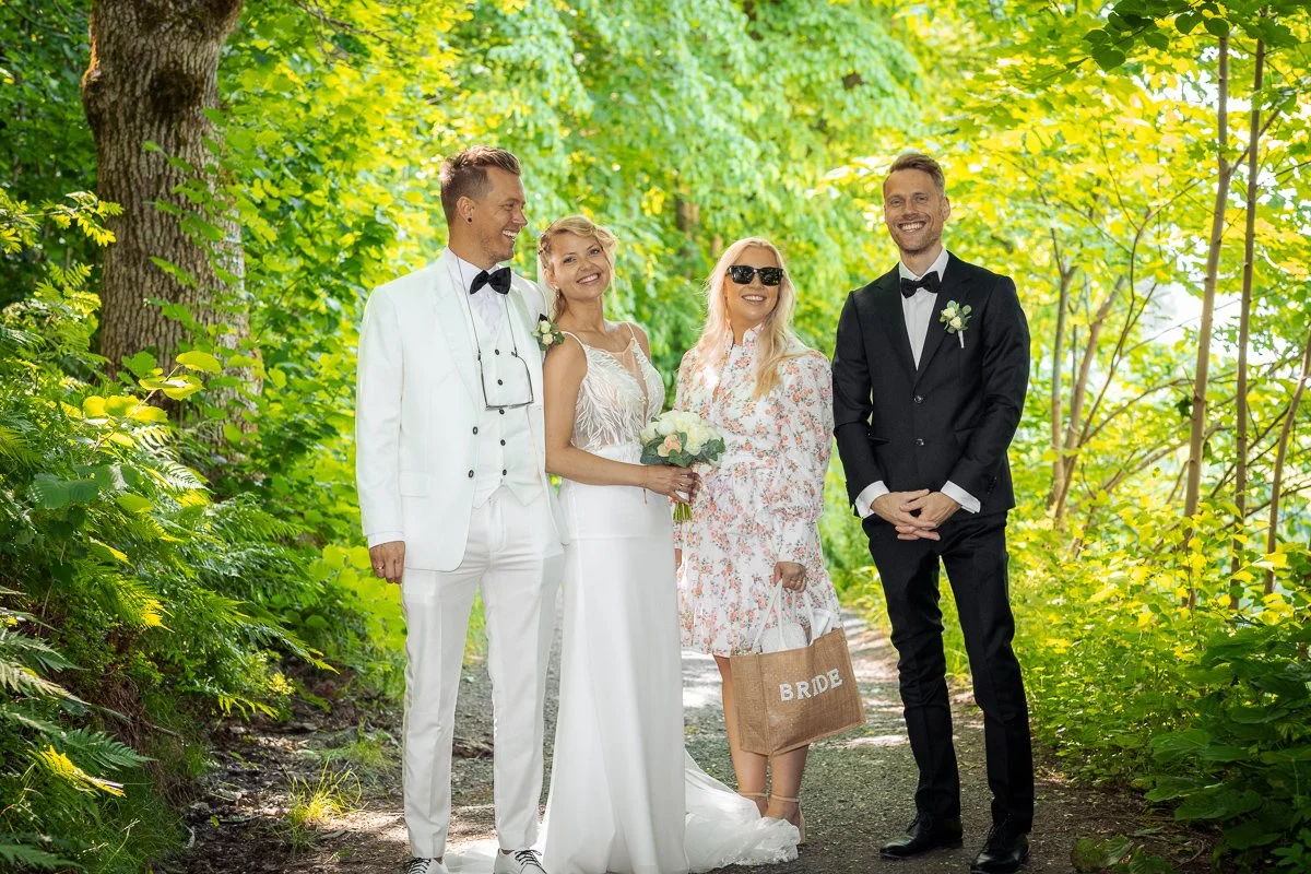En gruppe på fem personer, to menn og tre kvinner, smiler og står på en skogssti under grønne trær, iført formelle klær, en av kvinner har brudekjole og en av mennene har smoking, tilsynelatende på bryllupsdagen.