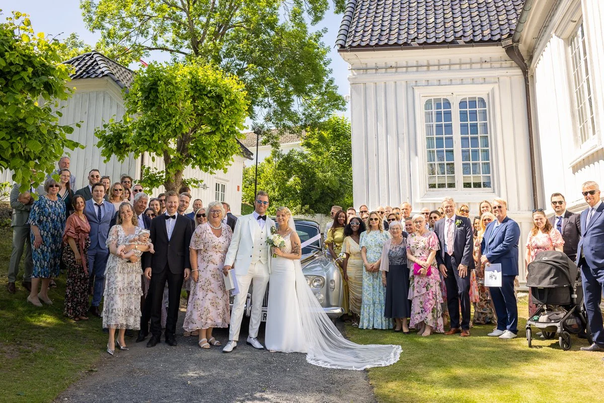 Bryllupsbilde av en gruppe mennesker utenfor en hvit bygning, med en brud og brudgom i midten, alle smilende og kledd i formelle klær.