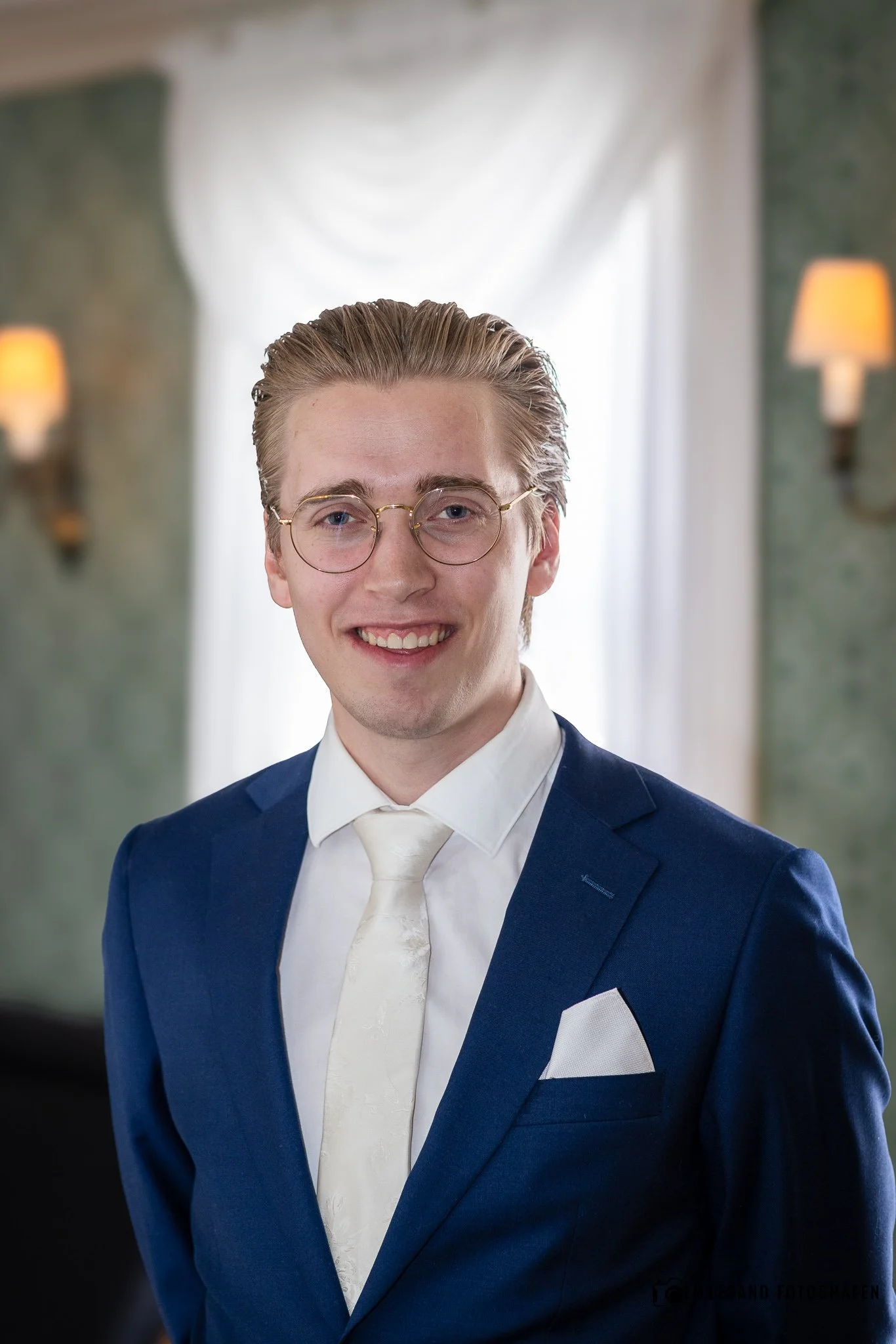 Portrett av en mann i formell dress med blå jakke, hvit skjorte og hvit slips, iført briller, smilende, innendørs med lyse gardiner og vegglamper