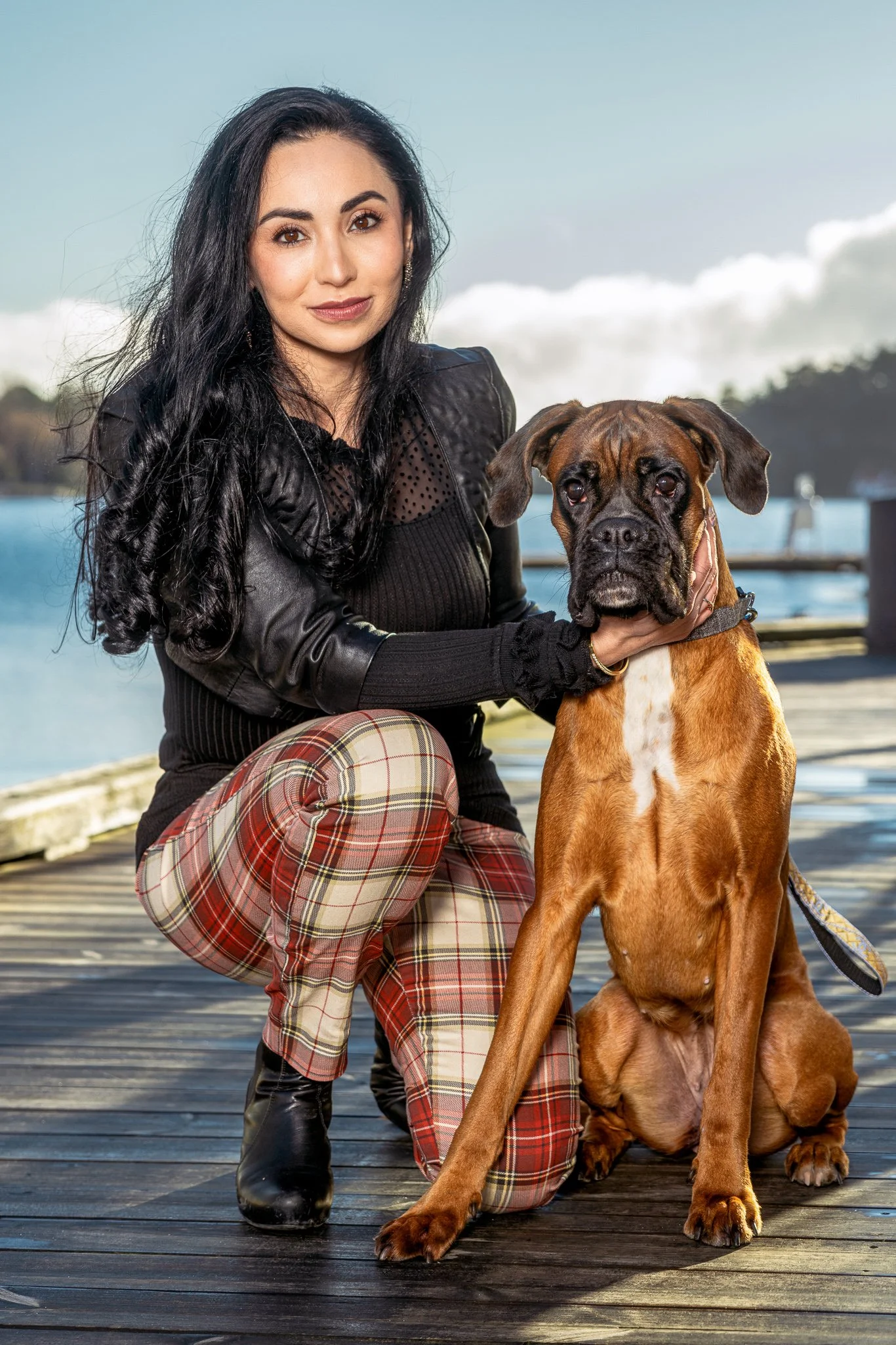 Dame med svart hår og svarte klær ved siden av stor hund på kai med vann og fjell i bakgrunnen.