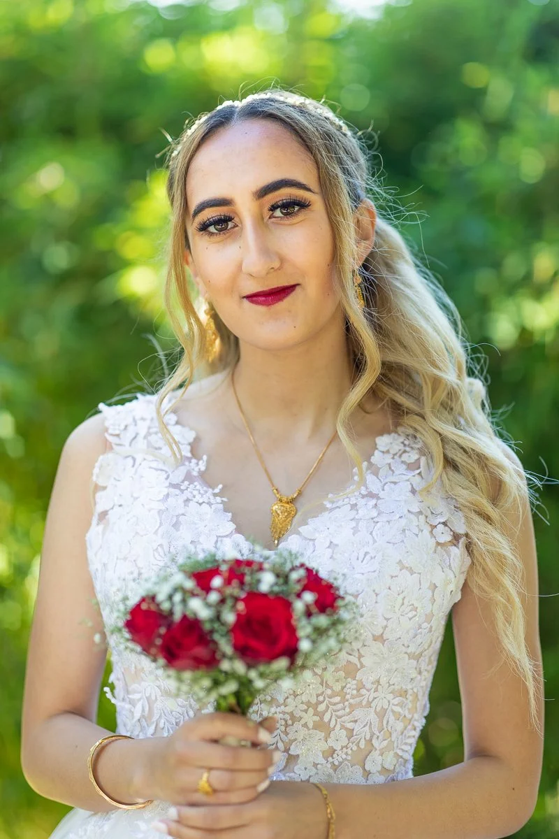 En kvinne i en brudekjole holder en bukett med røde blomster, utendørs med grønt løvverk i bakgrunnen.