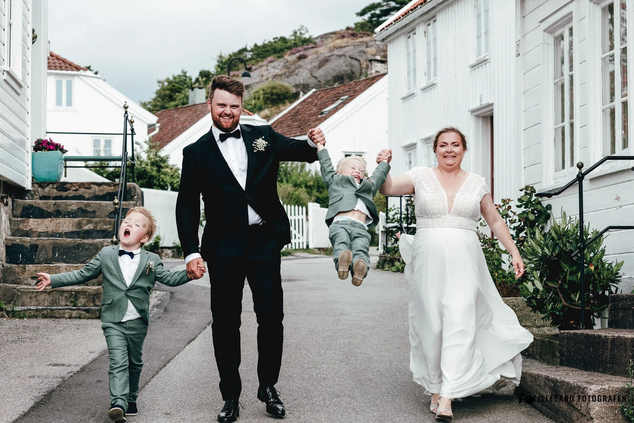 En lykkelig familie går i bryllupsklær på en gate. Mann i svart dress og kvinne i hvit kjole holder hender med to barn i dress. Barnet i midten hopper opp og familie smiler.