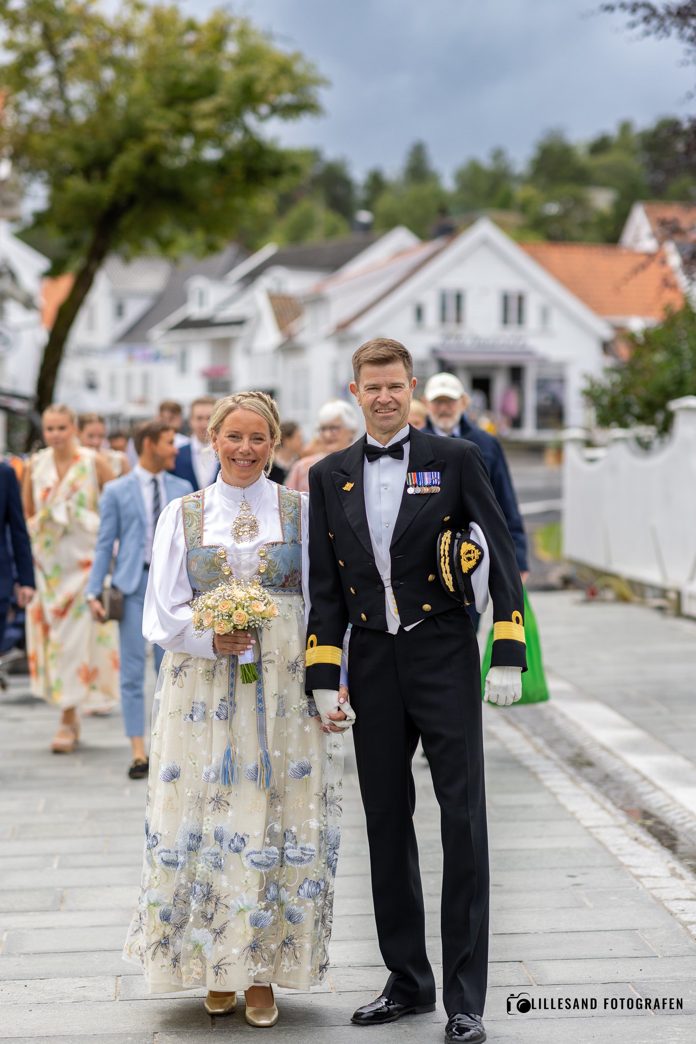 Et par går hånd i hånd under en bryllupsseremoni på en gate med hvite hus, omgitt av familie og venner. Kvinnen bærer en tradisjonell brudekjole og holder en blomsterbuket, mens mannen er i militæruniform.
