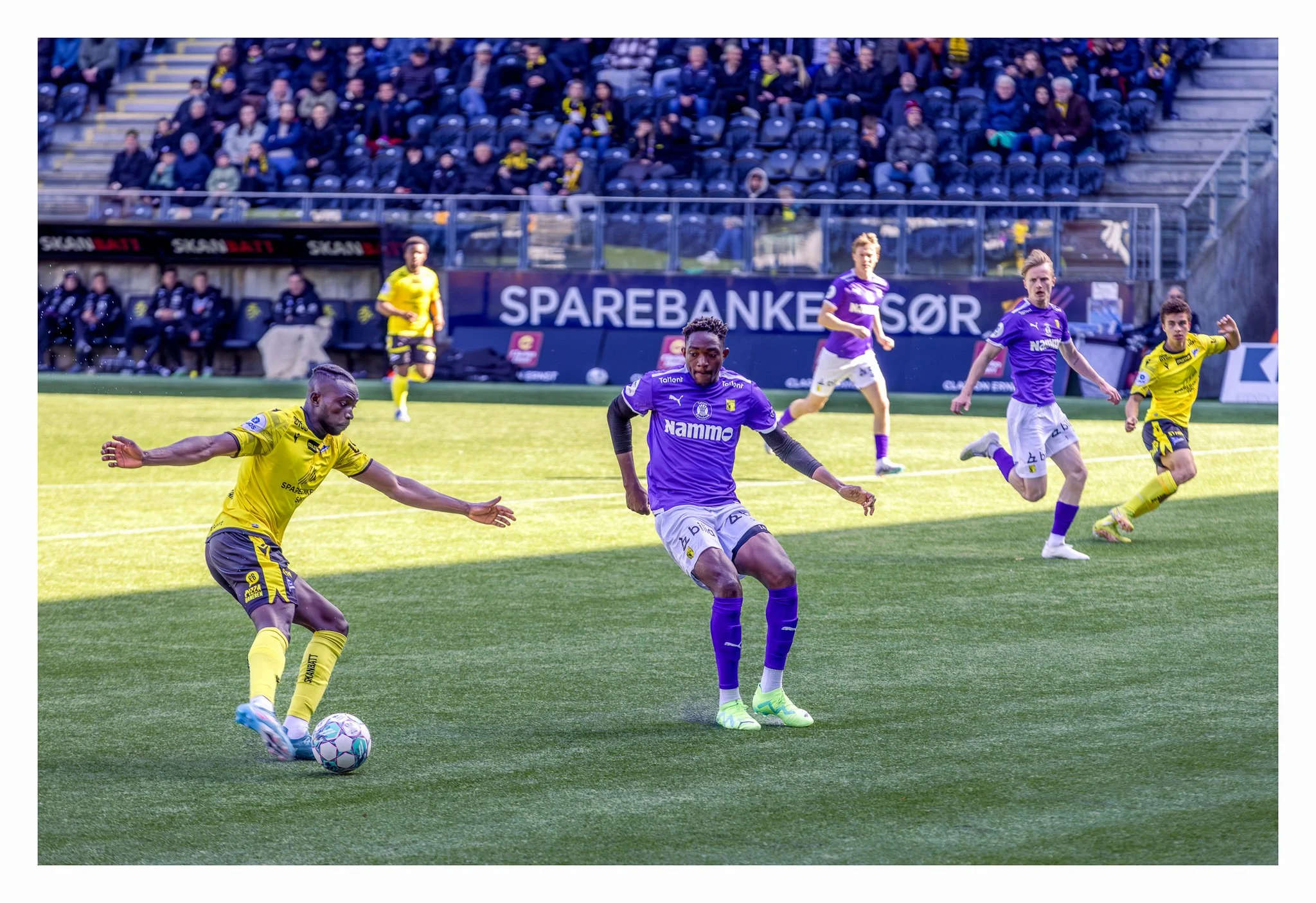 Fotballspillere i kamp, med noen i gule og andre i lilla trøyer, på banen under en kamp med publikummere i bakgrunnen.