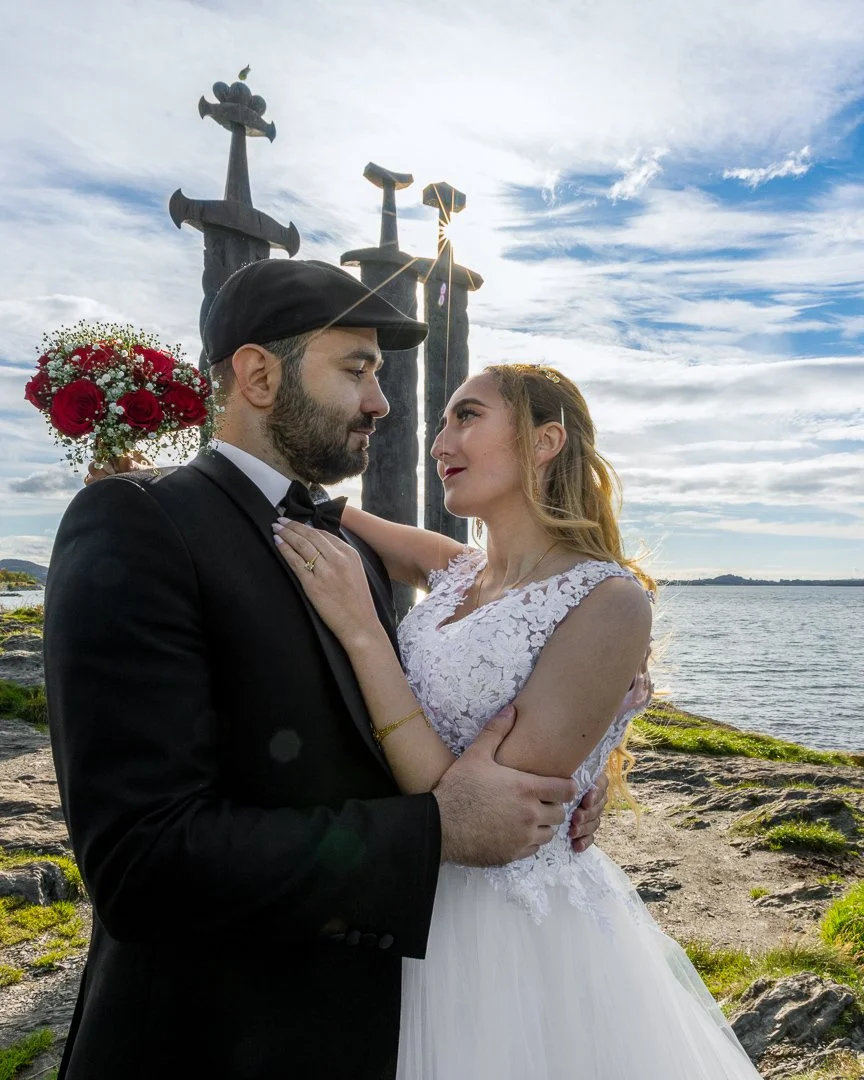 Et brudepar står ansikt til ansikt på en steinstrand, med en stor vannkropp og himmel med skyer i bakgrunnen. Bruden har på seg en hvit bordudekjole og har langt, blond hår. Brudgommen har på seg smoking og en svart lue. Det er tre monumentale sverds