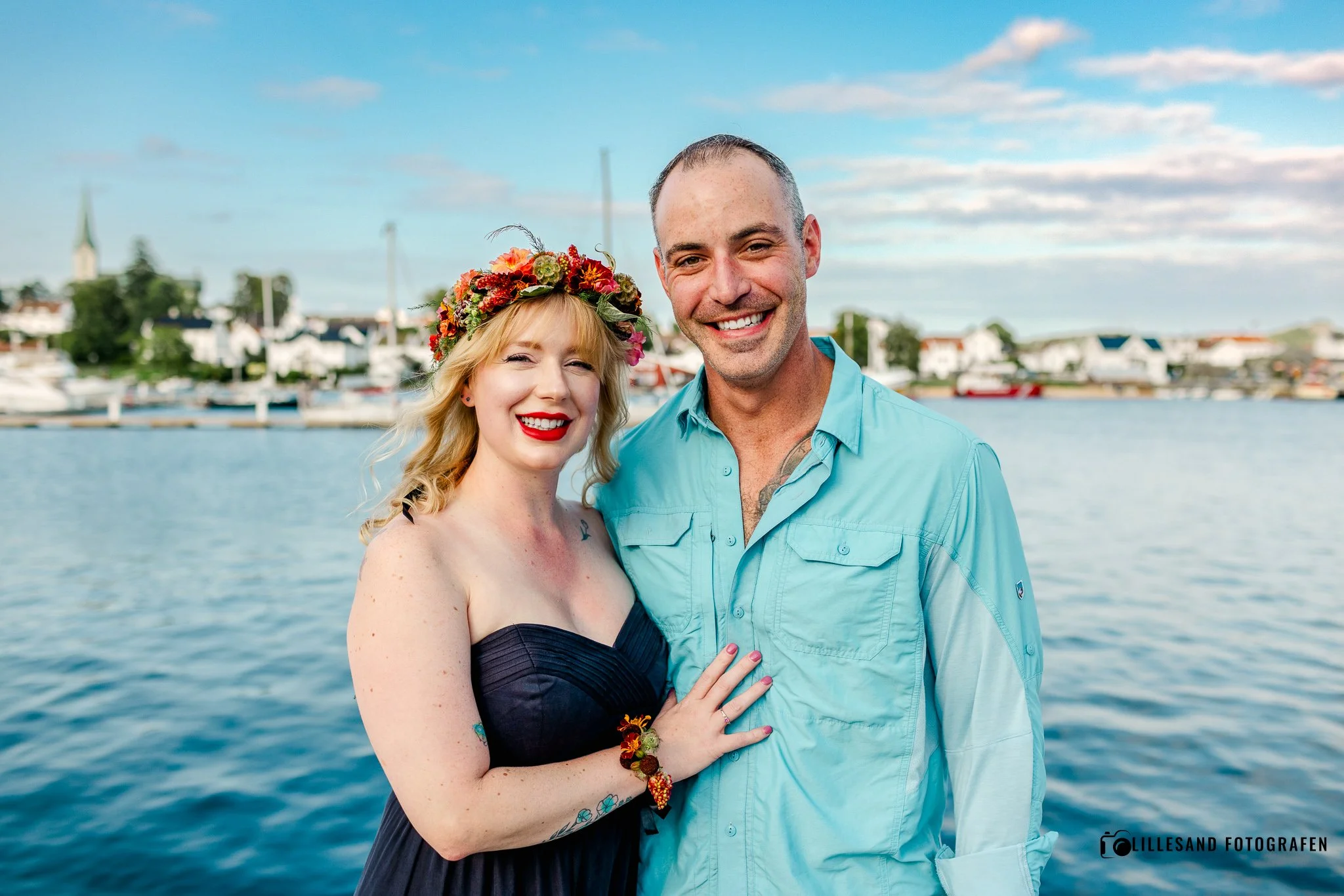 Et smilende par foran vann med båter og by i bakgrunnen, kvinnen har blomsterkrans og rødt lepper, mannen i blå skjorte.