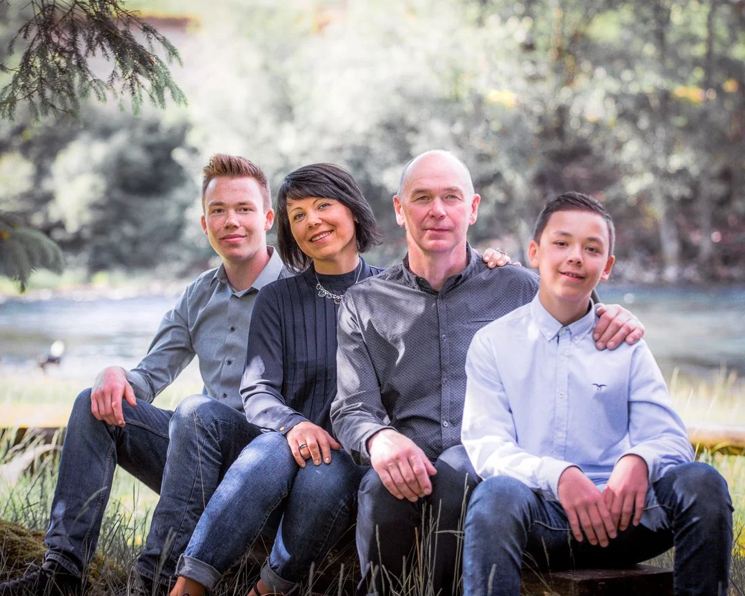 En familie på fire som sitter på en stokk ved en elv, omgitt av trær i et skogsområde.