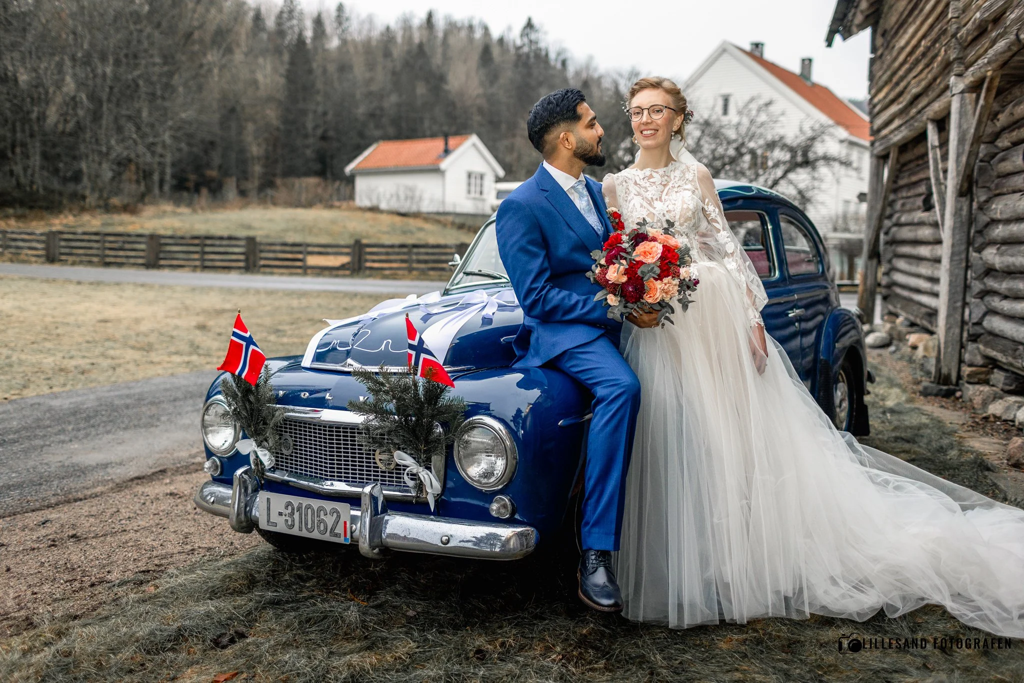 Et er par i brudekjole og dress som sitter på panseret på en blå vintage bil, pyntet med norske flagg og julegrener, foran en landlig bolig med trær i bakgrunnen.